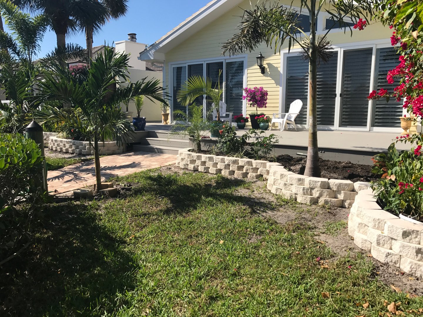 Tropical garden with palm trees and flowering plants in front of a yellow house with large windows and a patio area.