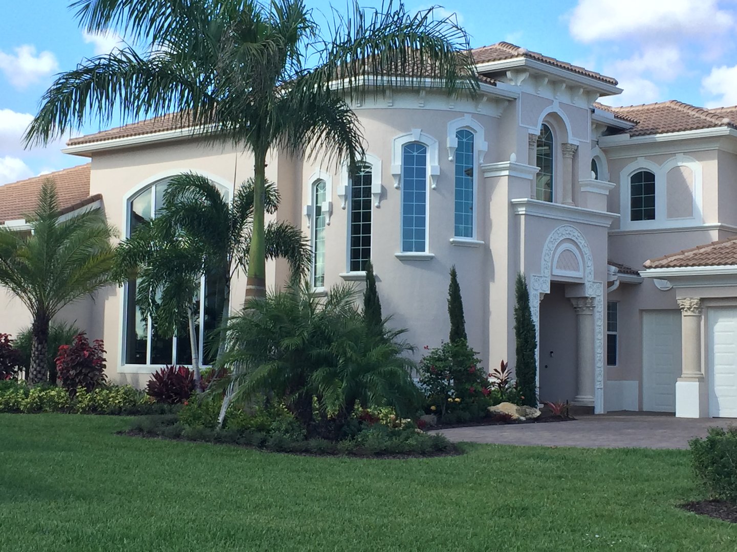 A large, modern Mediterranean-style house with arched windows, palm trees, and a well-manicured lawn under a blue sky.