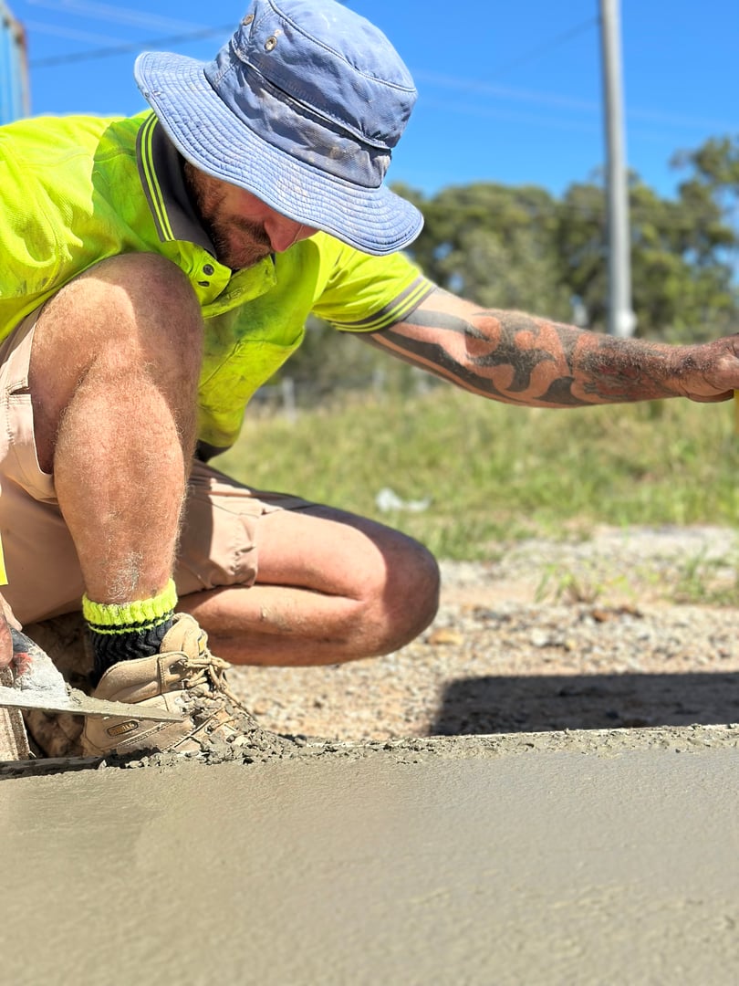 Worker in a yellow shirt and blue hat smoothing wet concrete with a trowel on a sunny day.
