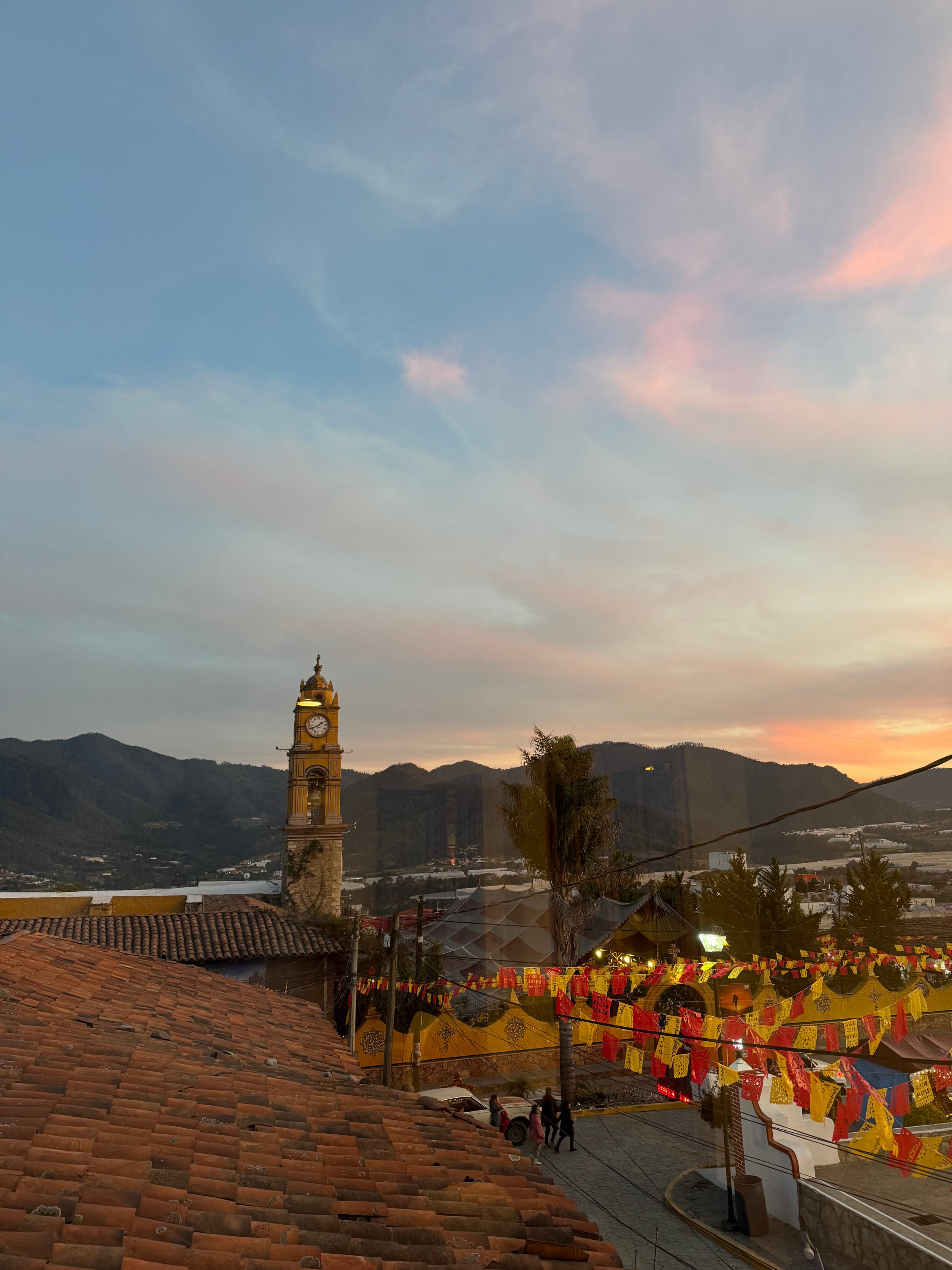 Clock tower and colorful banners at sunset in a town with mountainous landscape in the background.