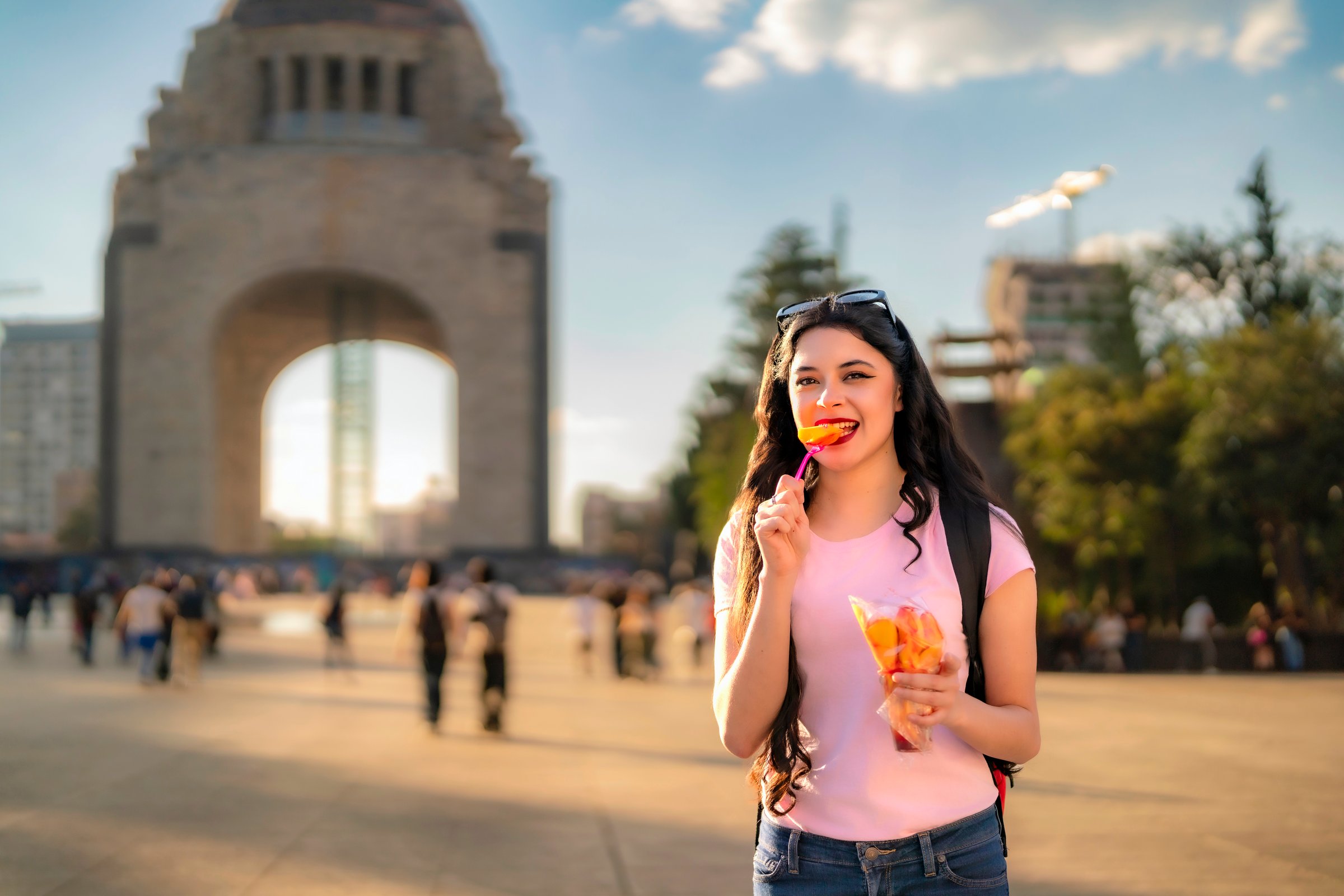 Smiling young Latina woman with wavy hair, pink t-shirt, jeans, and a red backpack enjoys spicy mango in front of the Monument to the Revolution in Mexico City during a sunny afternoon.