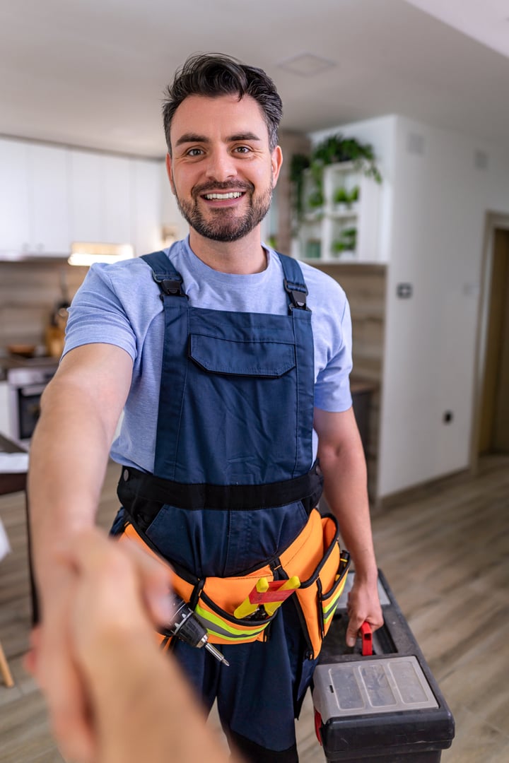 Handyman with tools smiles and offers handshake in a bright, modern kitchen environment.