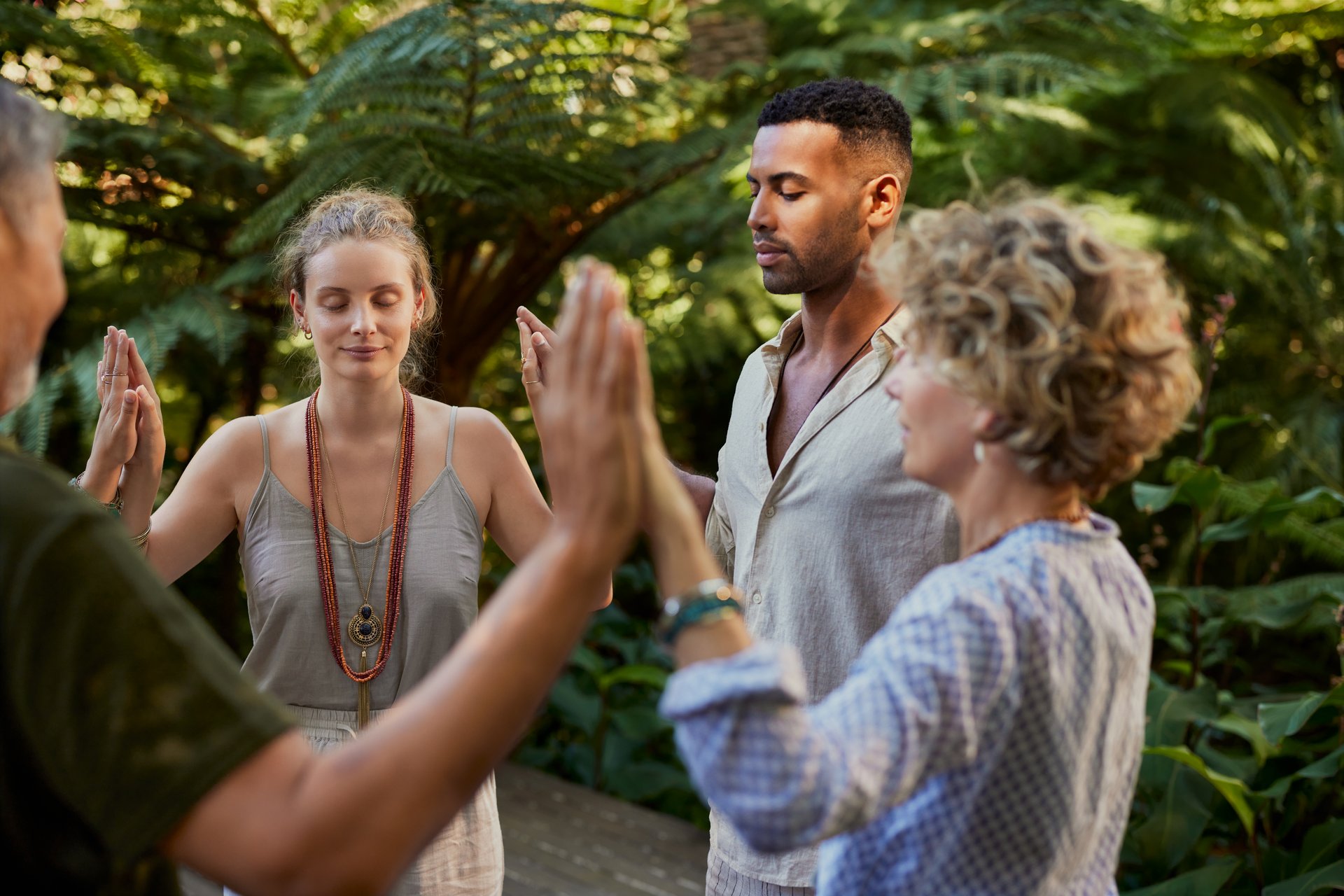 Group of diverse people stand in a circle holding hands, sharing a healing and spritual moment in nature. Multiethnic people at wellness retreat connect through energy practice in a lush outdoor setting. Adults meditate and praying together, creating a sense of unity and calm.