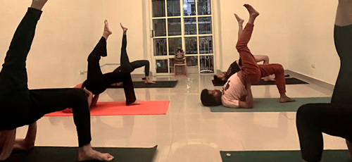 Person lying on a yoga bolster on a mat, with exercise equipment like a roller, weights, and blankets in the background.