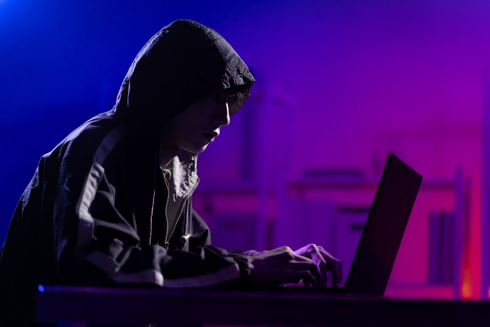 A young man in a black hoodie intently using a laptop in a neon-lit room, evoking themes of cybersecurity, hacking, coding, and digital privacy in a tech-driven world.