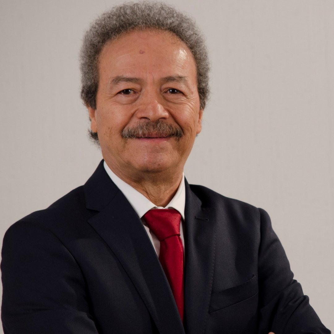 Smiling man with curly hair in a dark suit and red tie, arms crossed against a neutral background.