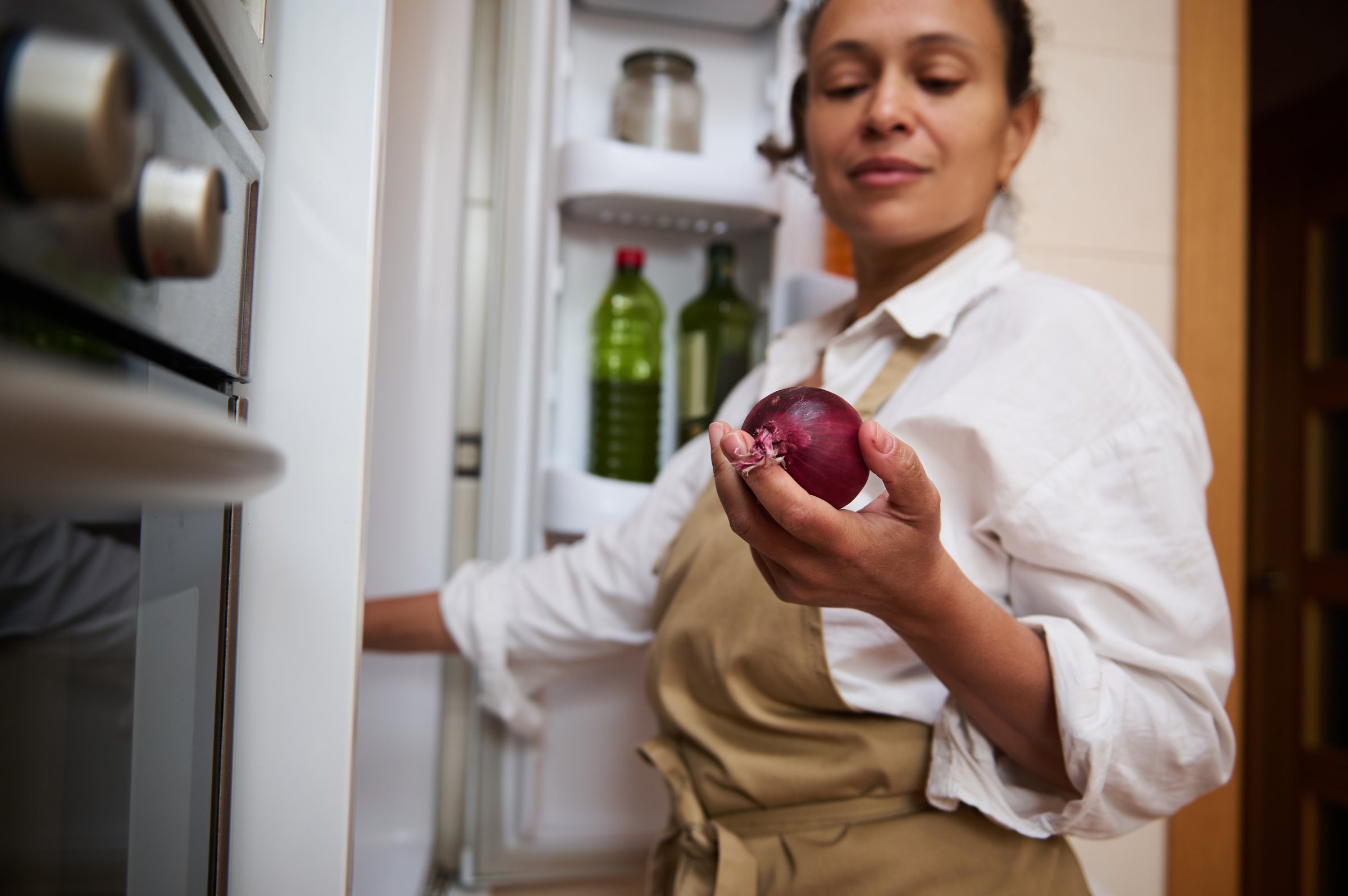 A woman in a kitchen apron holds a red onion near an open refrigerator, preparing ingredients for cooking.