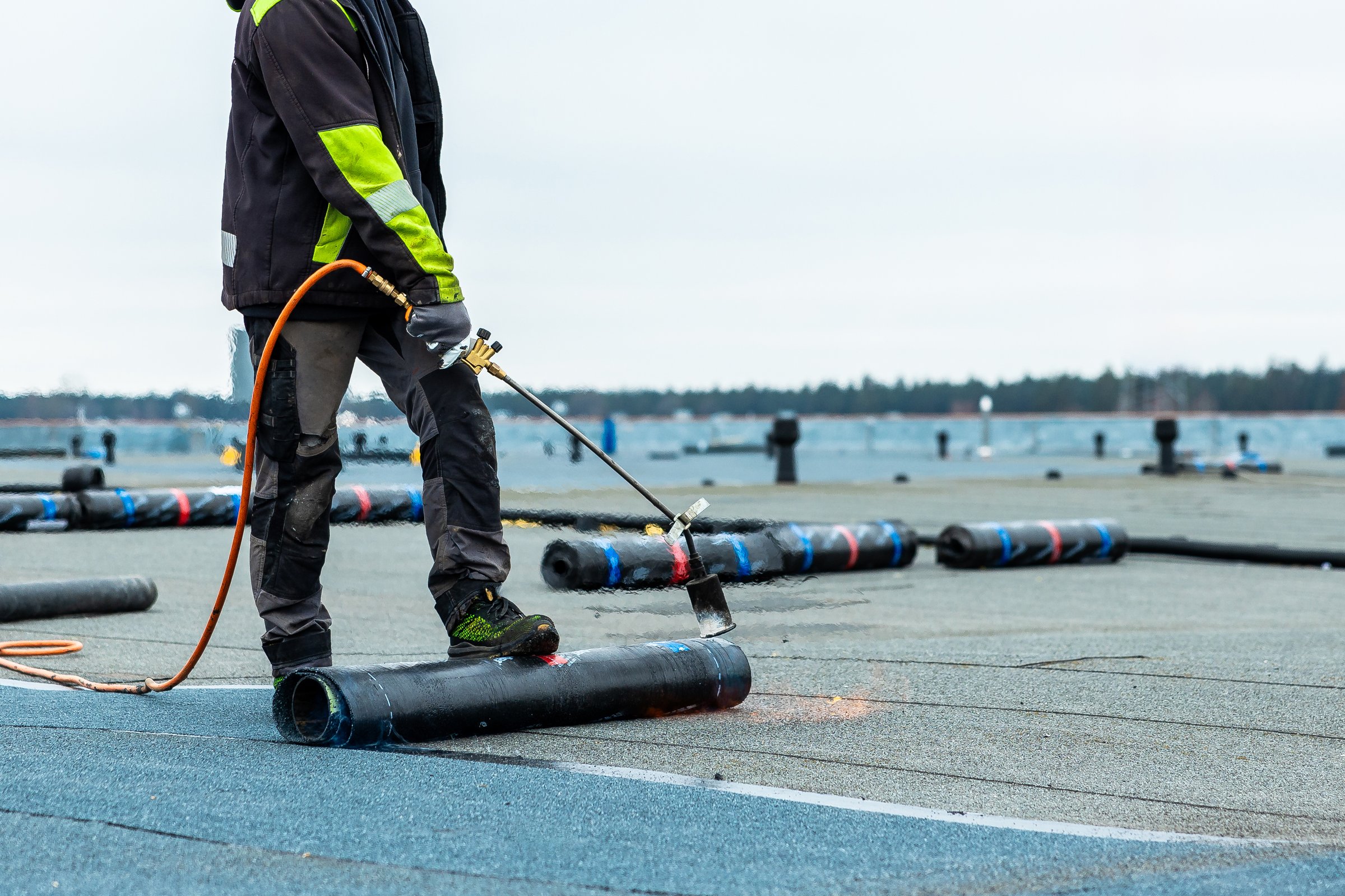 A worker in protective clothing uses a flame torch to apply roofing material on a flat rooftop. A tree lined shore and body of water are visible nearby.