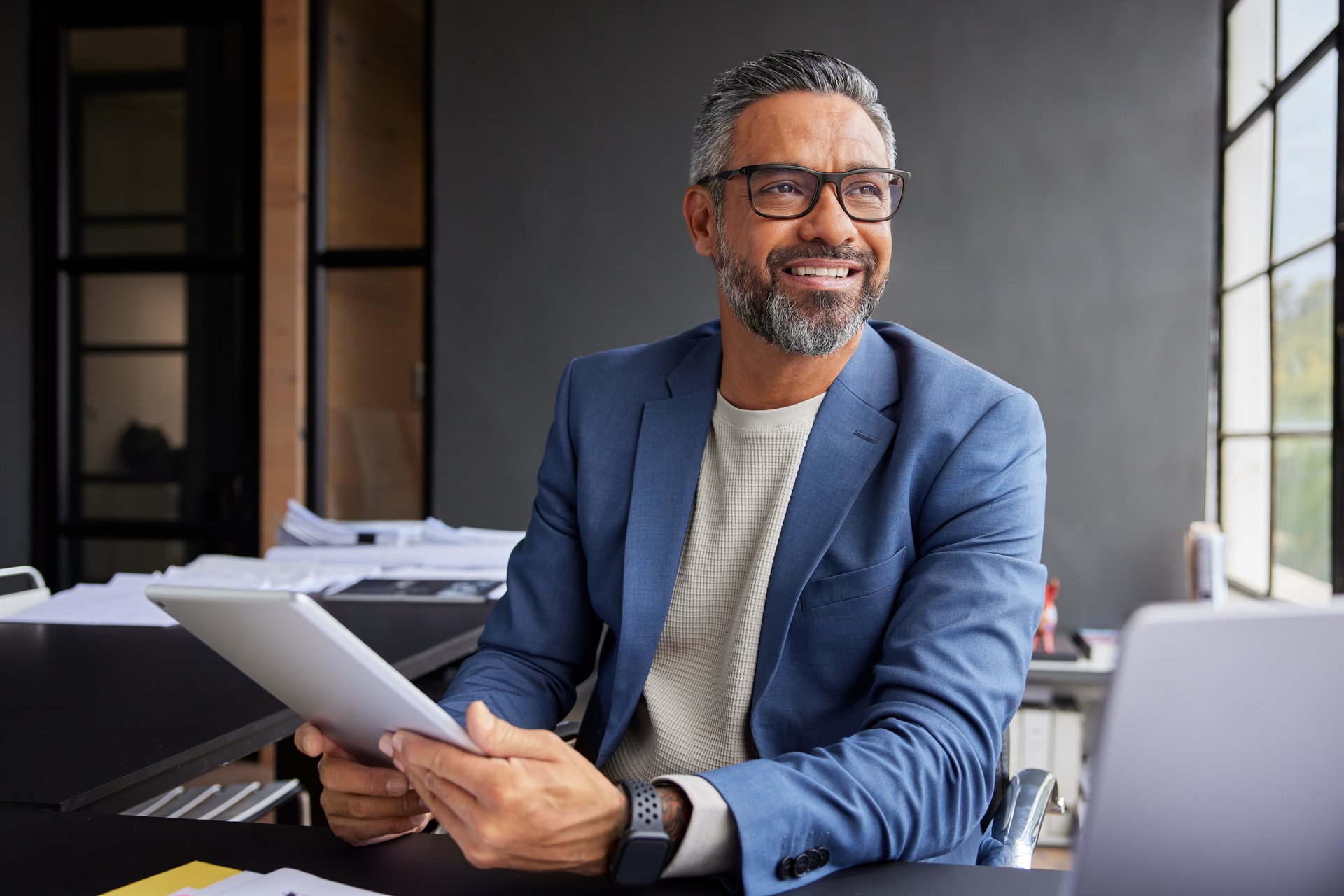 Cheerful mixed race businessman holding a tablet, looking thoughtful in modern office. Professional middle eastern man engaged with tablet, smiling while gazing out of window. Confident business executive working with a digital tablet, daydreaming and aspiration concept.