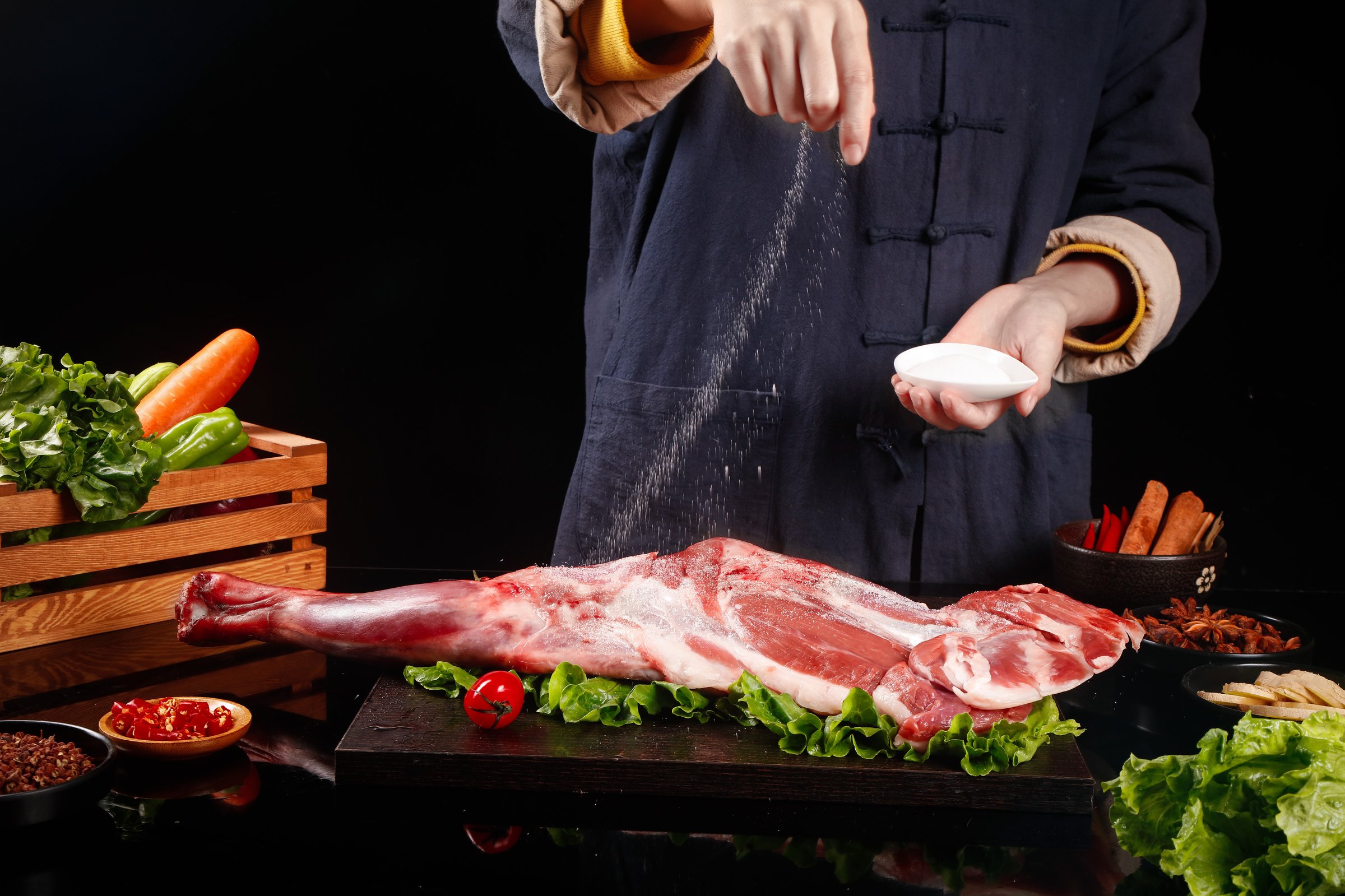 Chef seasoning raw lamb leg with salt on wooden cutting board surrounded by fresh vegetables and spices in professional kitchen setting