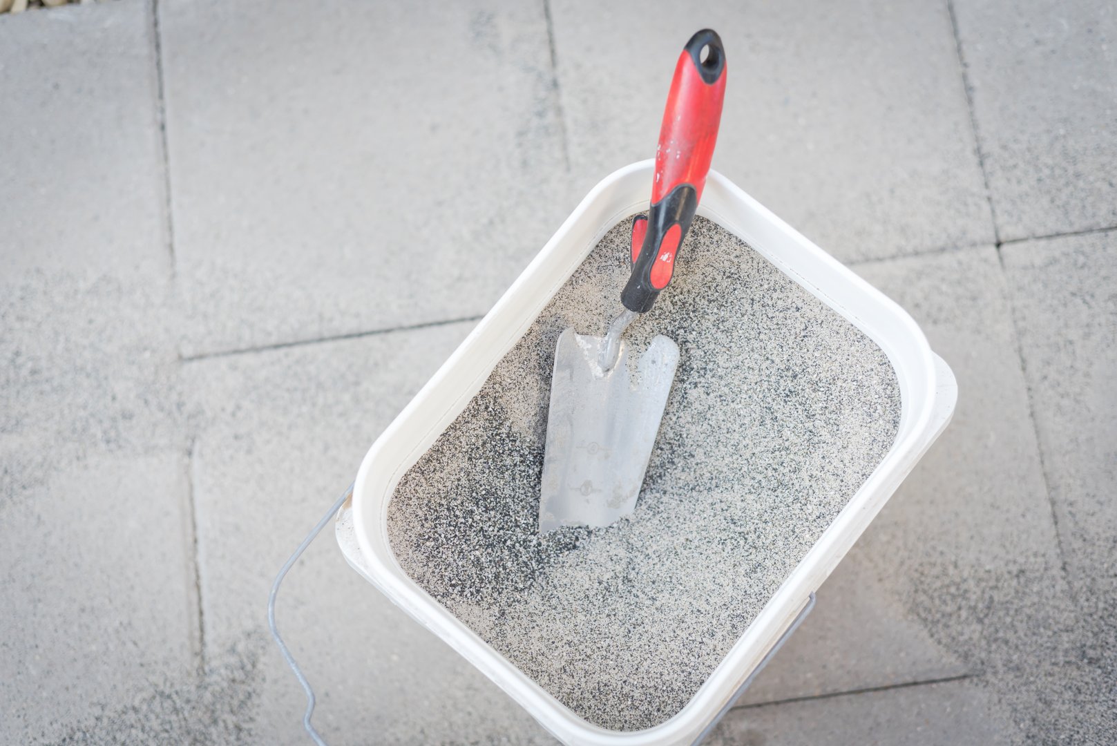 Full bucket of polymeric paver sand with trowel on top of gray concrete patio stone paver walkway. This material to fill joint between pavers to firm and prevent weeds from growing up between