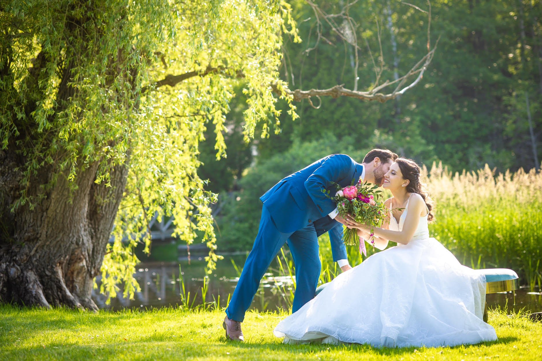 Bride and groom kissing by a pond under a tree, with the groom leaning and holding a bouquet, on a sunny day.