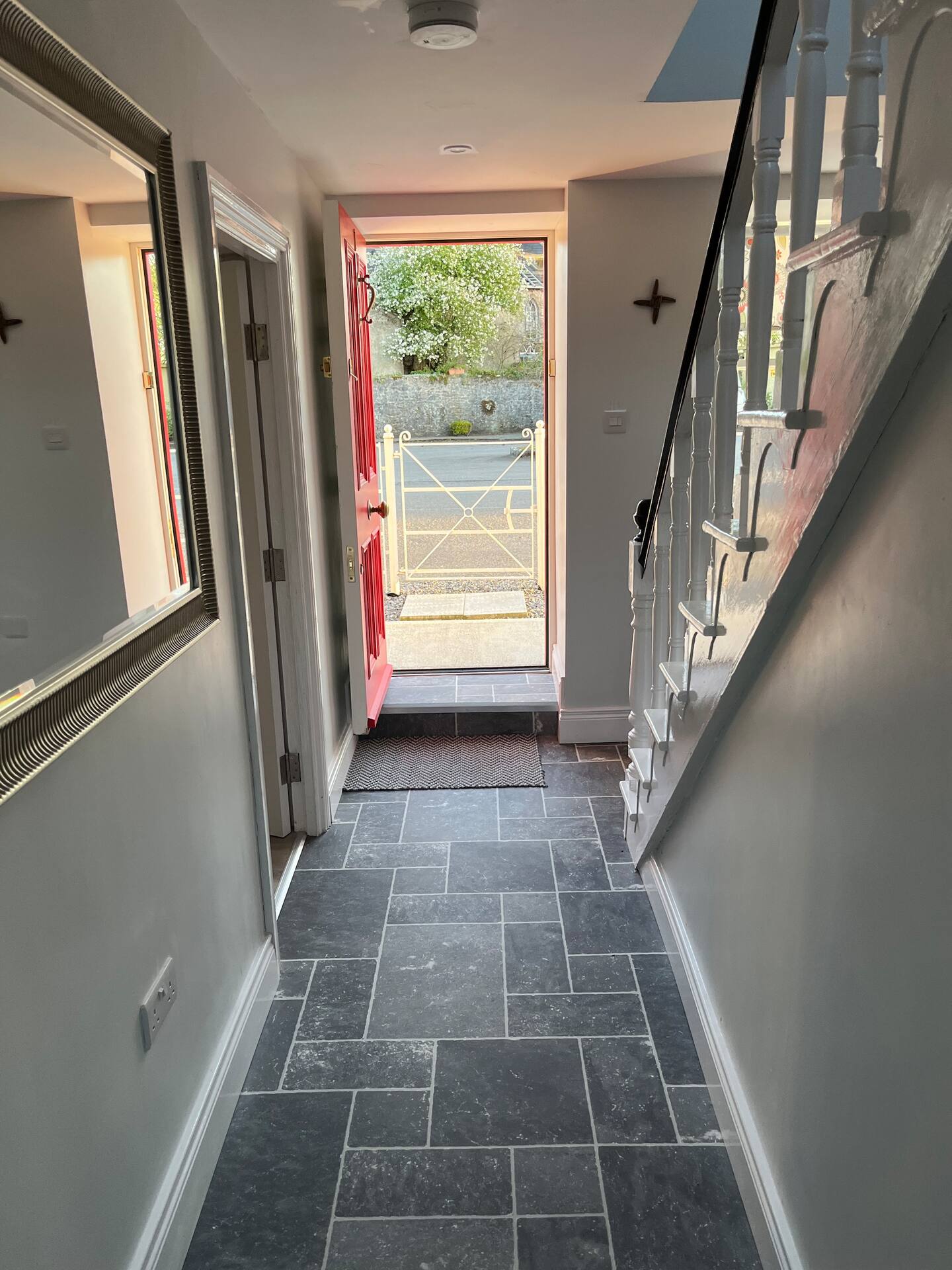 Hallway with gray tiled floor, open red front door, staircase on the right, and a mirror on the left wall.