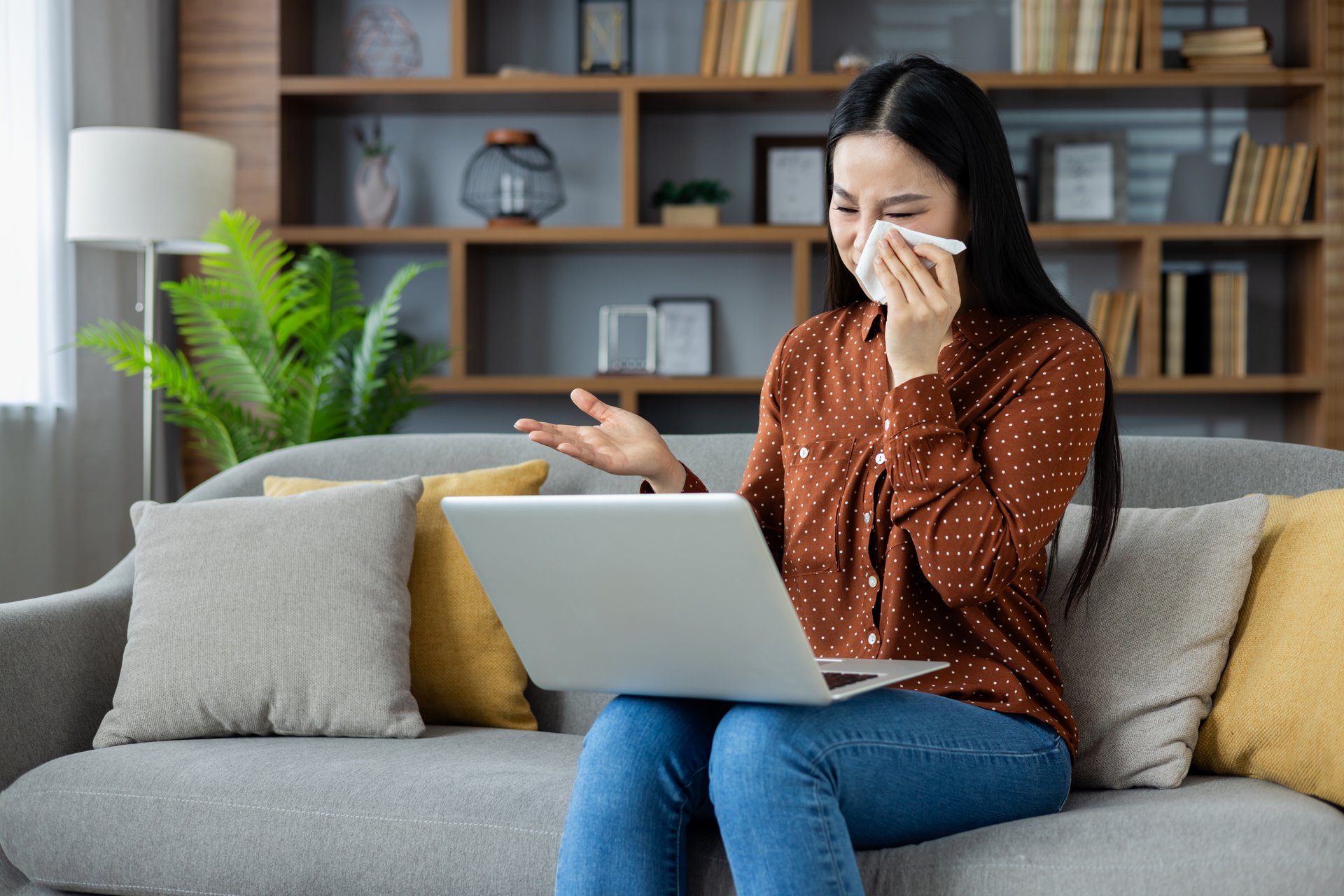 Asian woman using laptop on sofa, holding tissue, appears emotional, possibly video chatting. Wearing casual attire, setting suggests home environment with emphasis on comfort and technology.