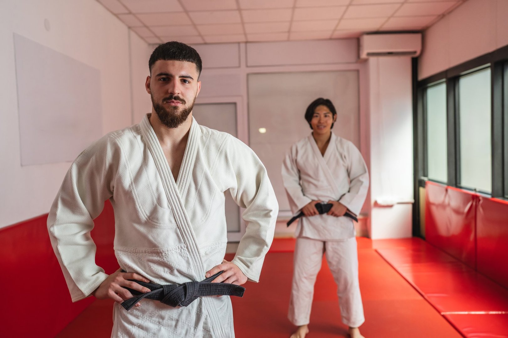 Two judo athletes, one in the foreground and one in the background, are posing in a kimono wearing white kimonos and black belts, showcasing their martial arts expertise