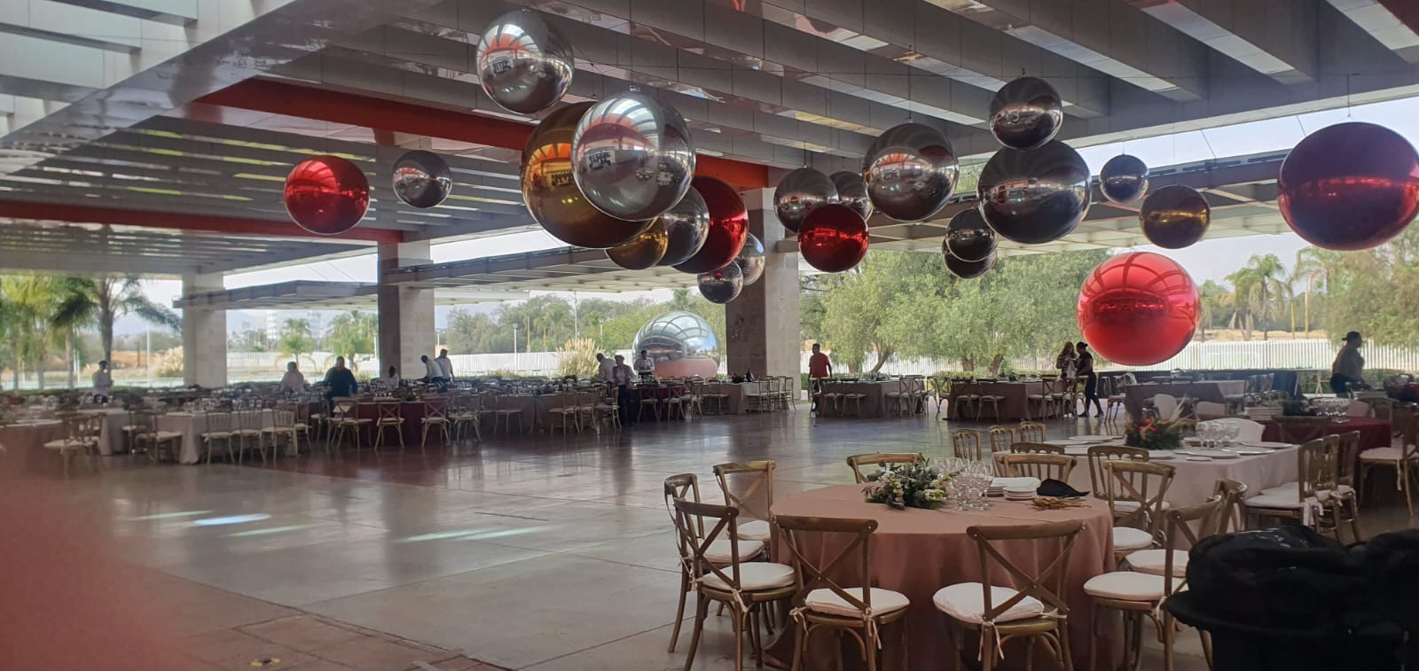 Large event venue decorated with red and silver balloons, tables set with white and pink tablecloths, lush greenery outside.