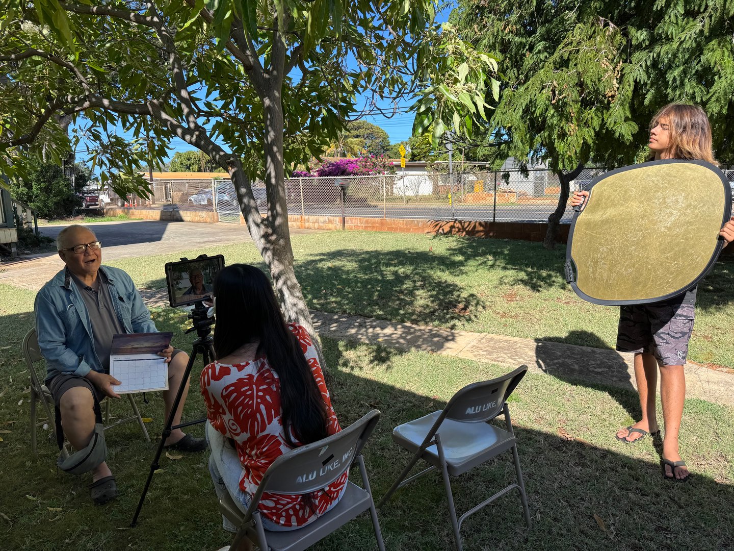 Outdoor interview setup with a man seated facing a camera on a tripod, a woman holding a tablet, and a person with a reflector.