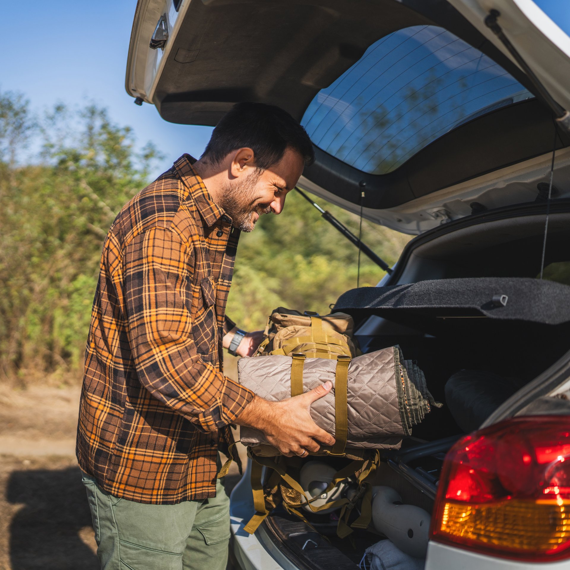 Adult man camper loads camping equipment into the trunk of a white car, surrounded by lush green scenery on a dirt road, ready for an outdoor adventure