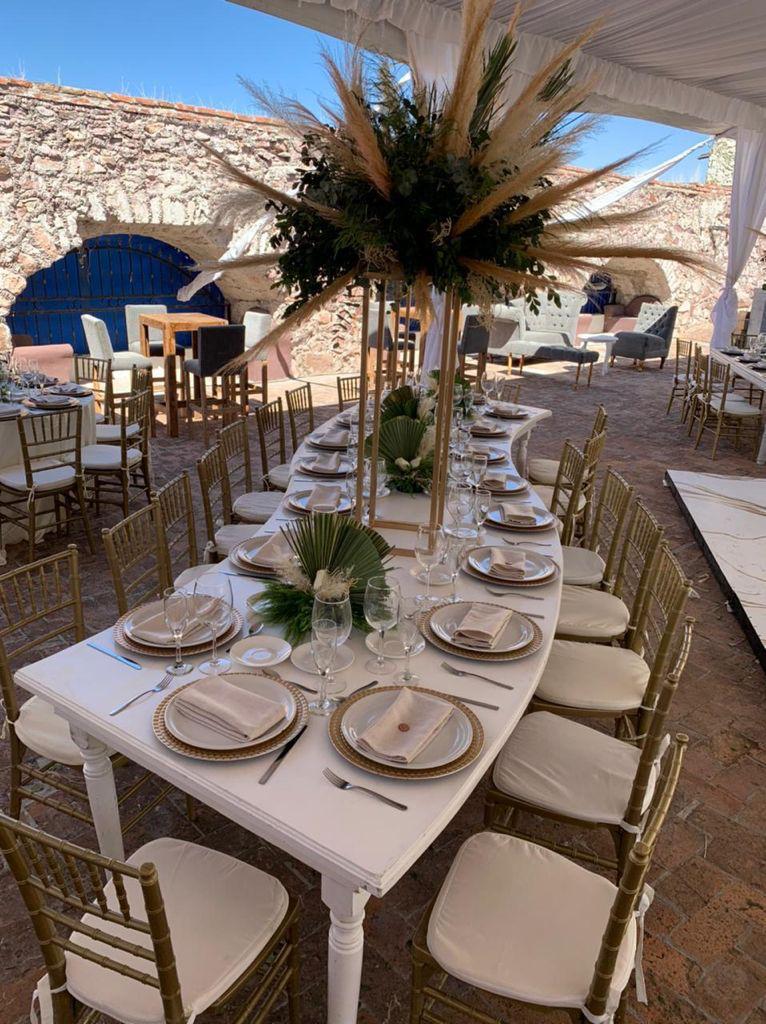 Elegant outdoor dining table setup with white plates, folded napkins, glassware, and a tall centerpiece of greenery and pampas.