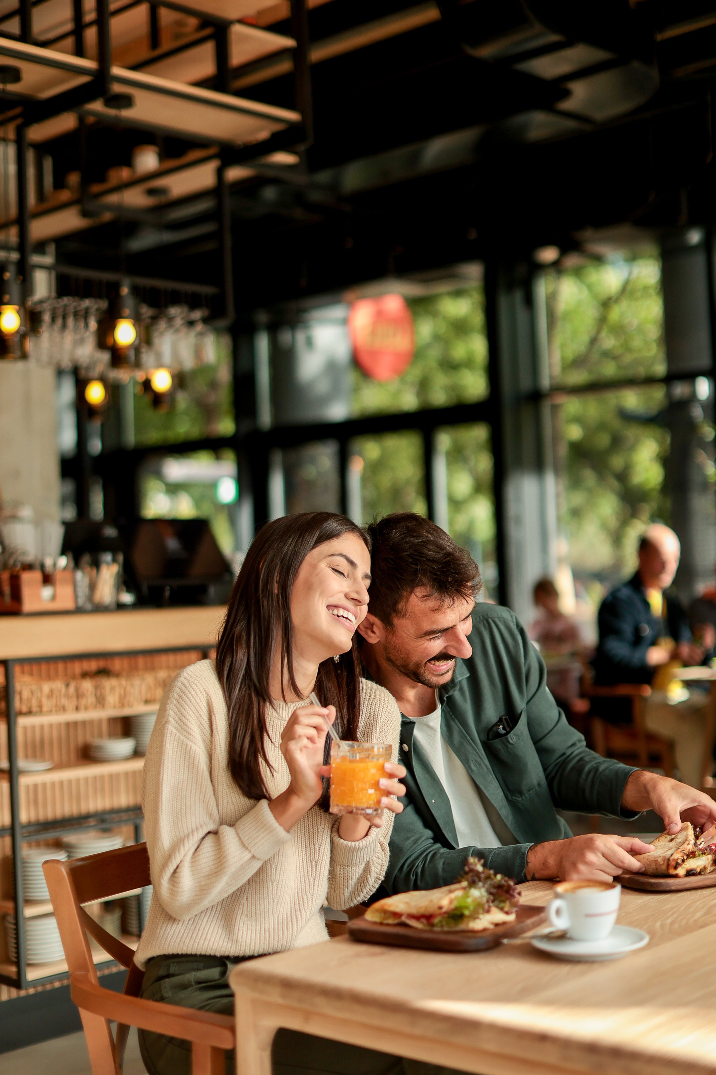 Couple shares laughter and delicious food while sitting at a wooden table in a bright cafe, surrounded by greenery and other patrons enjoying their meals