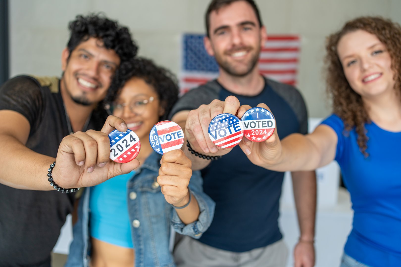 diverse group of smiling friends holding 2024 Vote election I voted buttons or pins at a US election polling station. In the background, american flag at the background, democracy in America USA