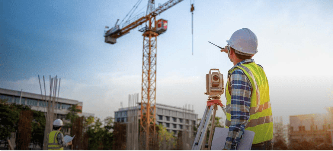 Construction worker using a theodolite at a site with a crane and building in the background, wearing a hard hat and vest.