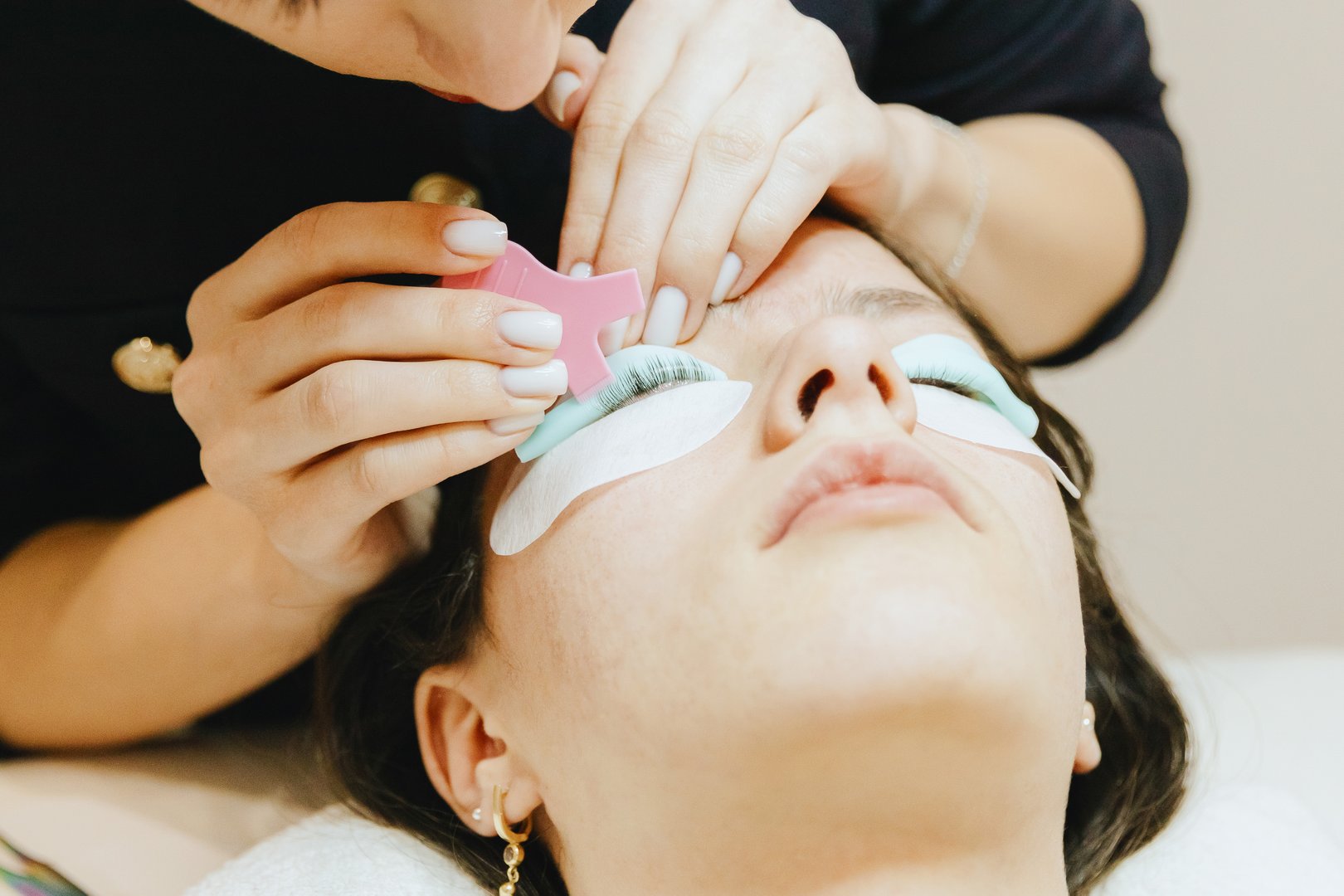 Portrait of a young beautiful Caucasian brunette cosmetologist girl in a dark jacket who combs, gluing eyelashes on the silicone roller of the left eye using a plastic brush to a teenage girl client lying on the cosmetology table in a home beauty salon for eyelash lifting, top side close-up view with selective focus .