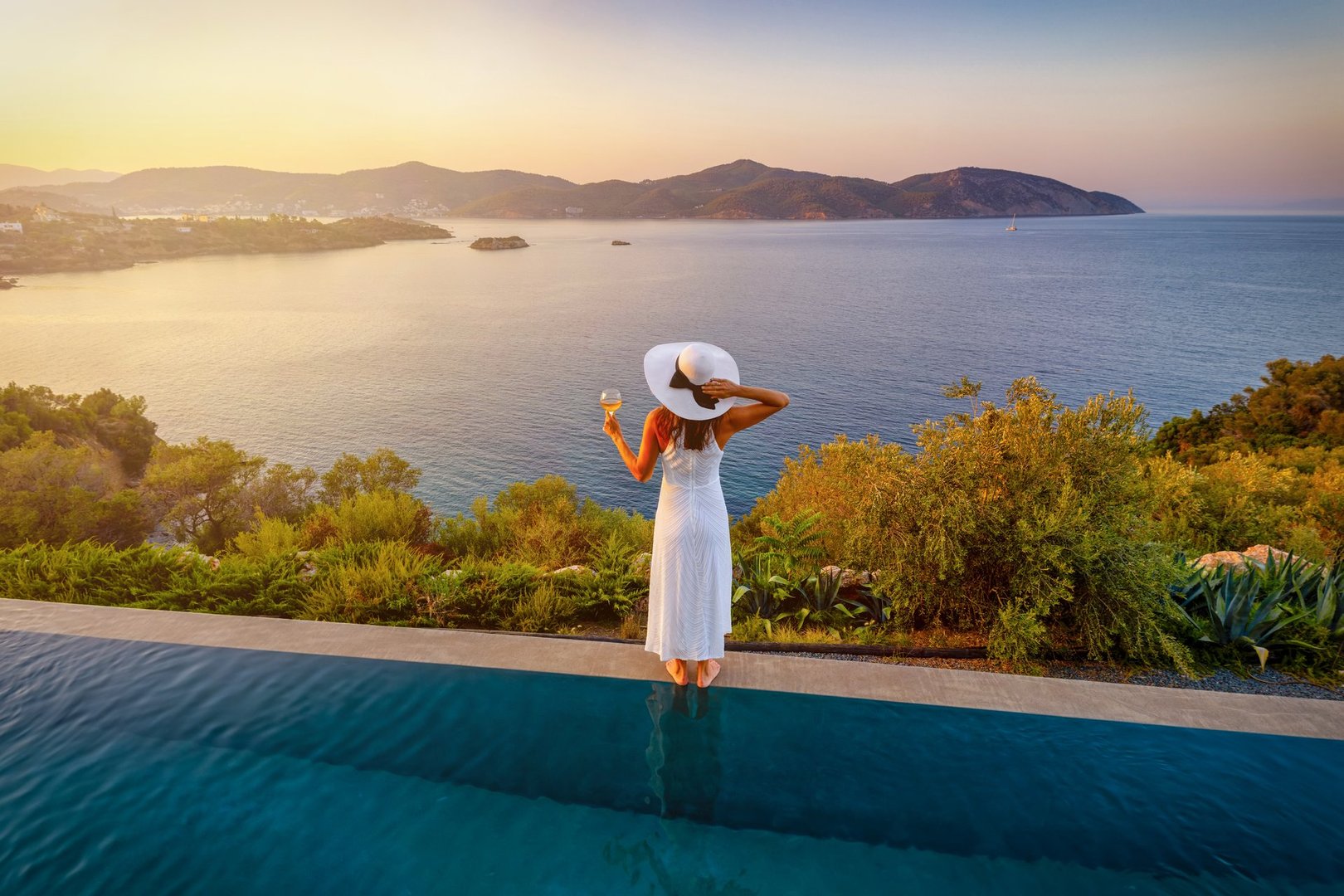 A elegant woman in a white dress stands by the swimming pool and enjoys the summer sunset behind the sea with a glass of wine