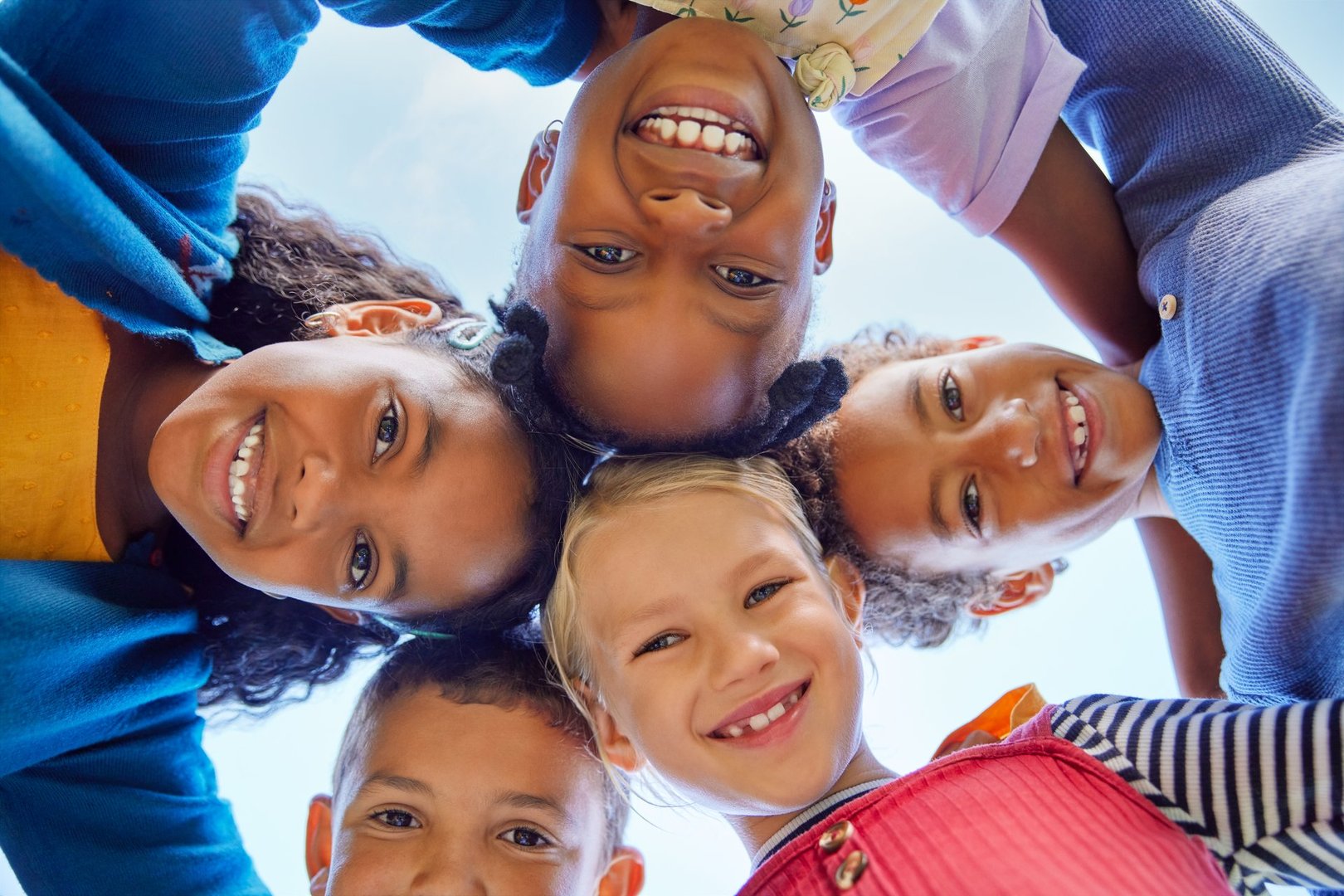 Low angle view of happy mixed race children embracing each other while smiling at camera. Cute little group of schoolchildren sticking heads together. Closeup face of cute girls and little boys embracing each other and smiling at camera while hugging.