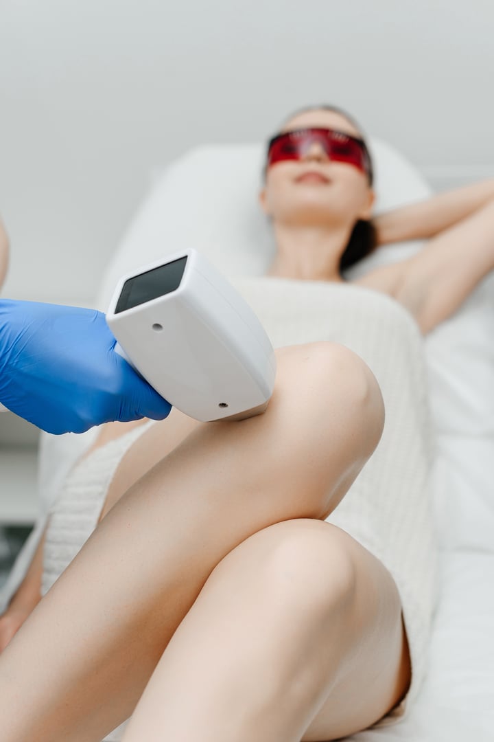 laser epilation, hair removal procedure, young beautiful caucasian smiling brunette woman lying on couch in beauty salon, legs epilation, red glasses, blue gloves, spa and cosmetology concept