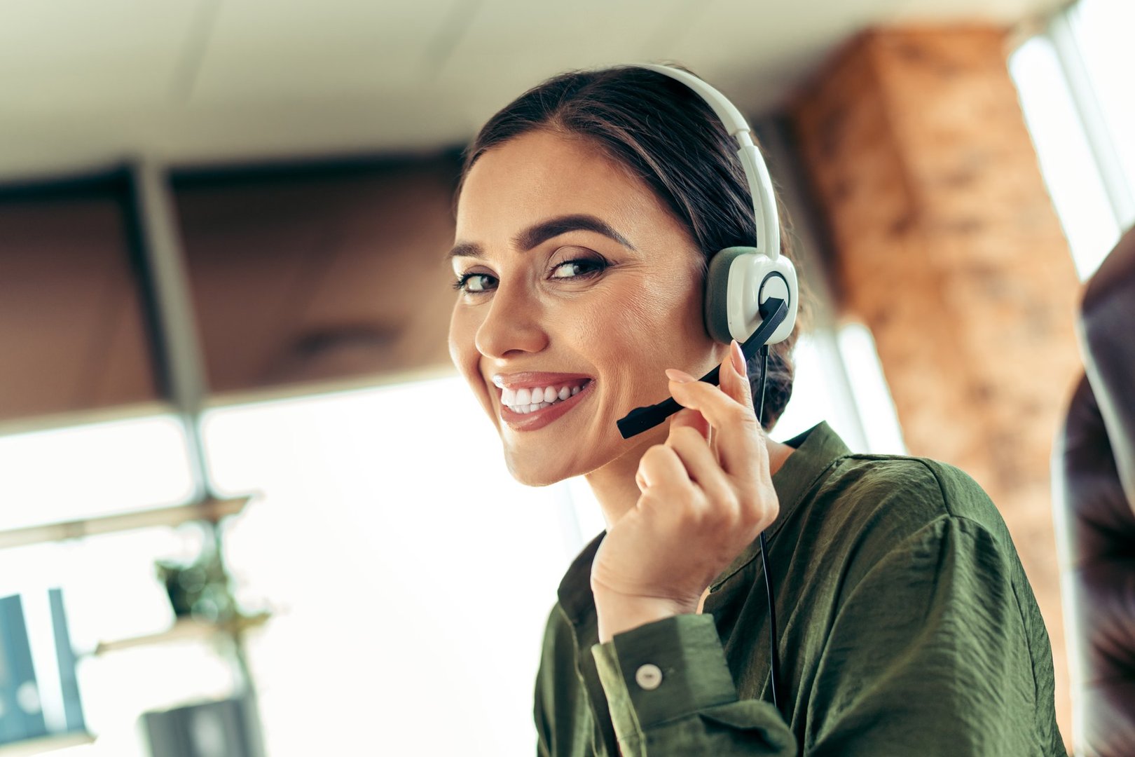 Smiling woman wearing a headset, working in a professional office setting, exuding confidence and offering excellent customer service with a friendly demeanor.