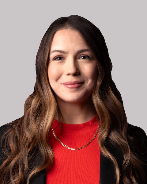 Woman with long brown hair, wearing a red top and black blazer, smiling softly against a grey background.