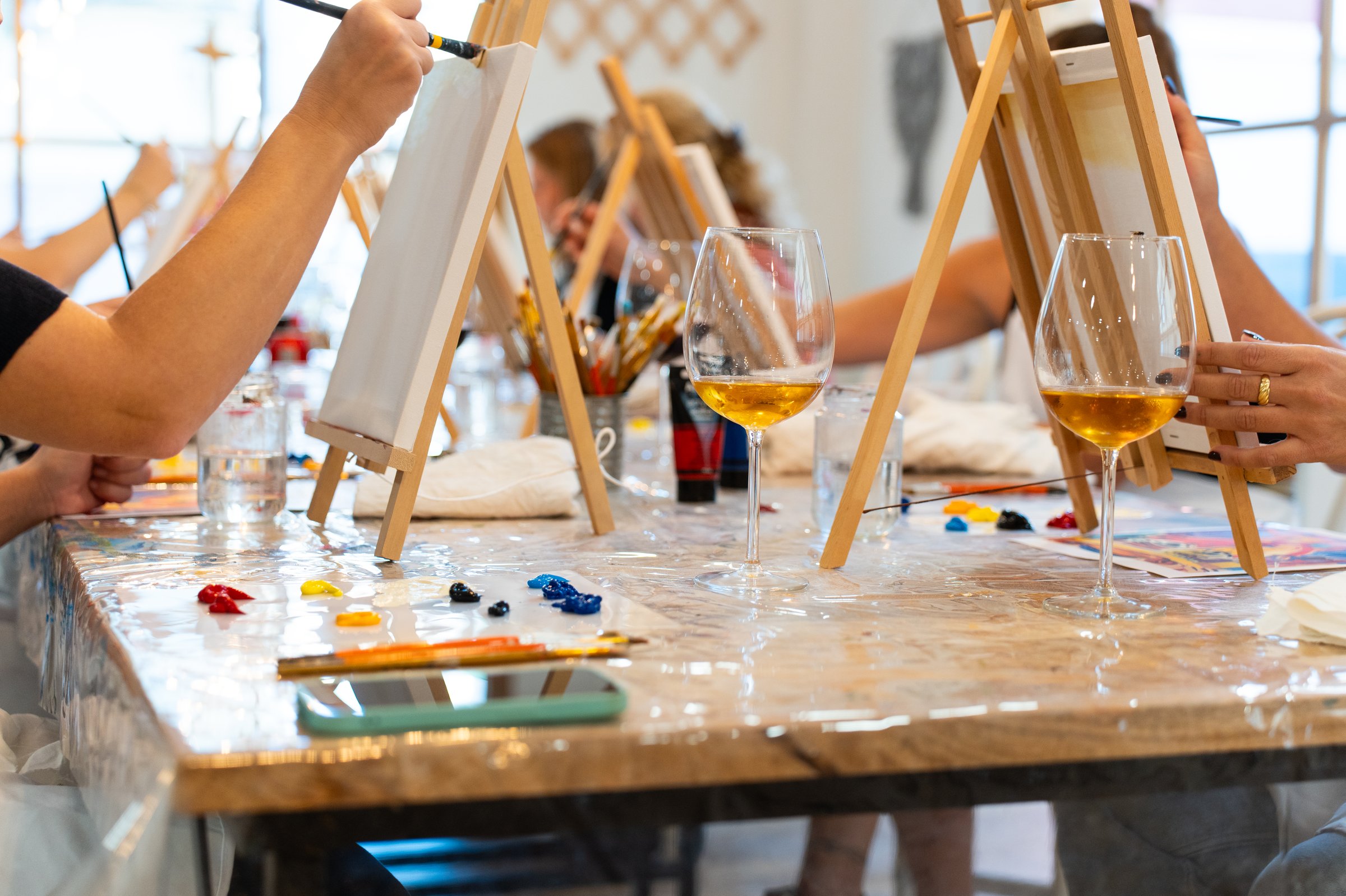 Group of friends painting on easels with wine glasses on the table, enjoying a creative and relaxing art class experience