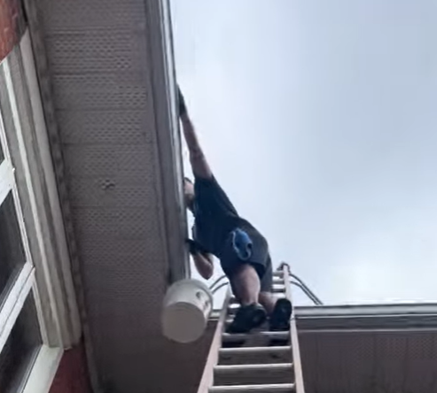 Person climbing a ladder to clean gutters on a house roof. A small bucket hangs from the ladder.