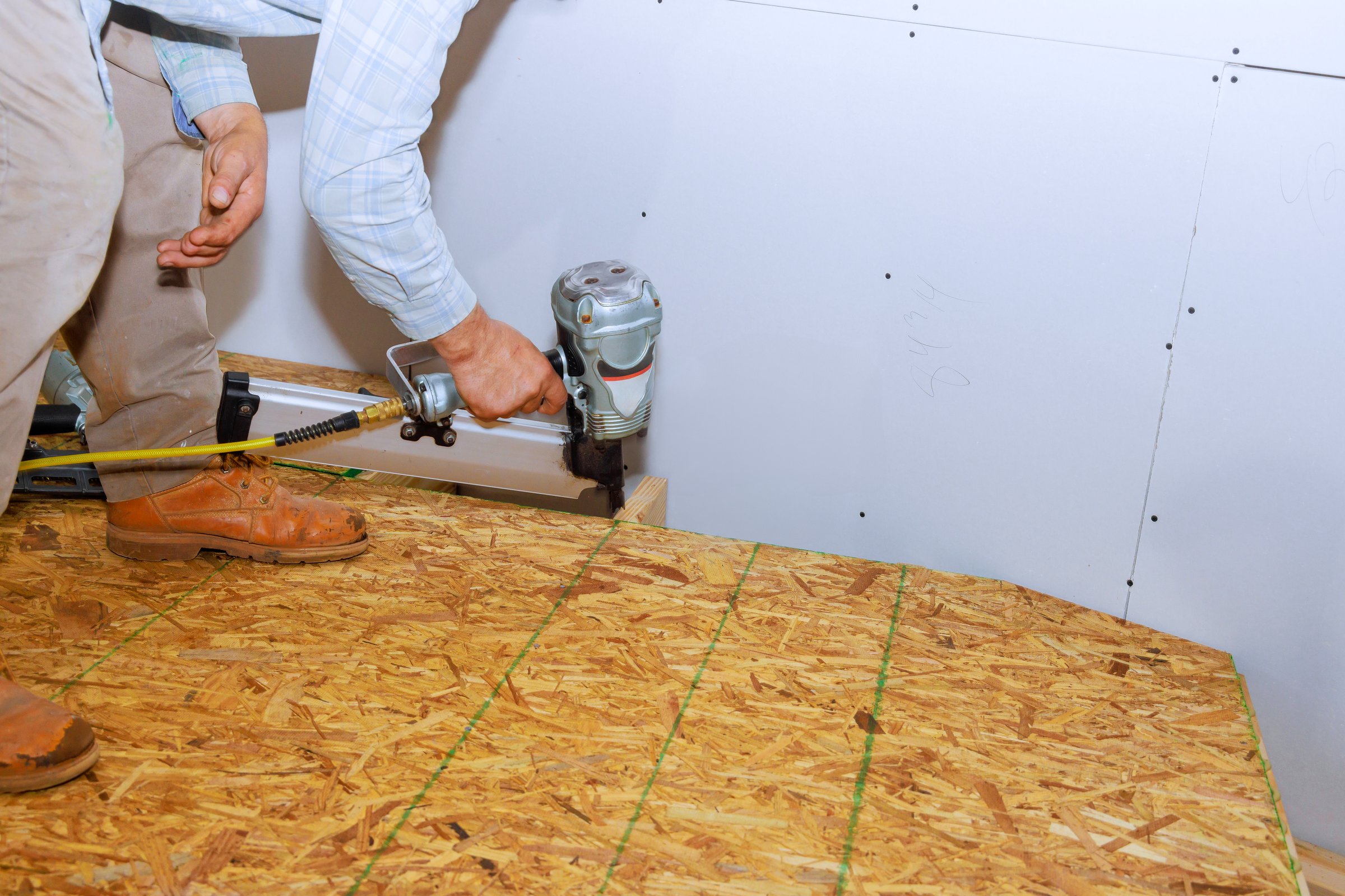 Construction worker kneels on OSB, using pneumatic nail gun to install flooring at job site.
