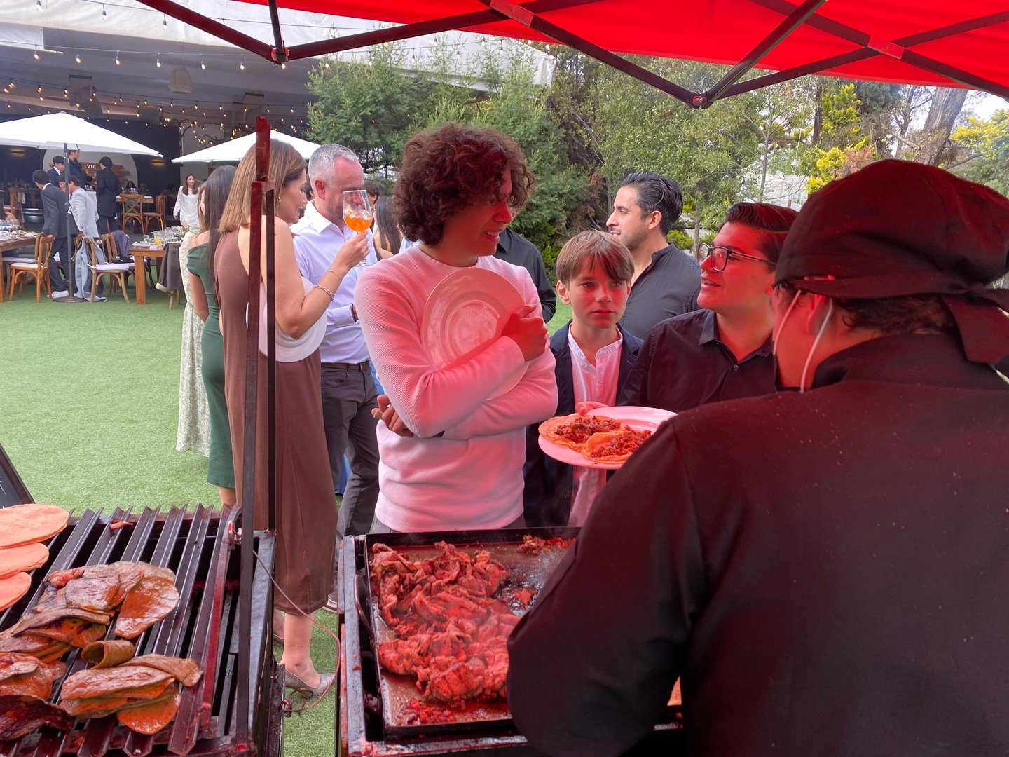 People gathered around a grill at an outdoor event, talking and serving food. Festive atmosphere with a red canopy overhead.