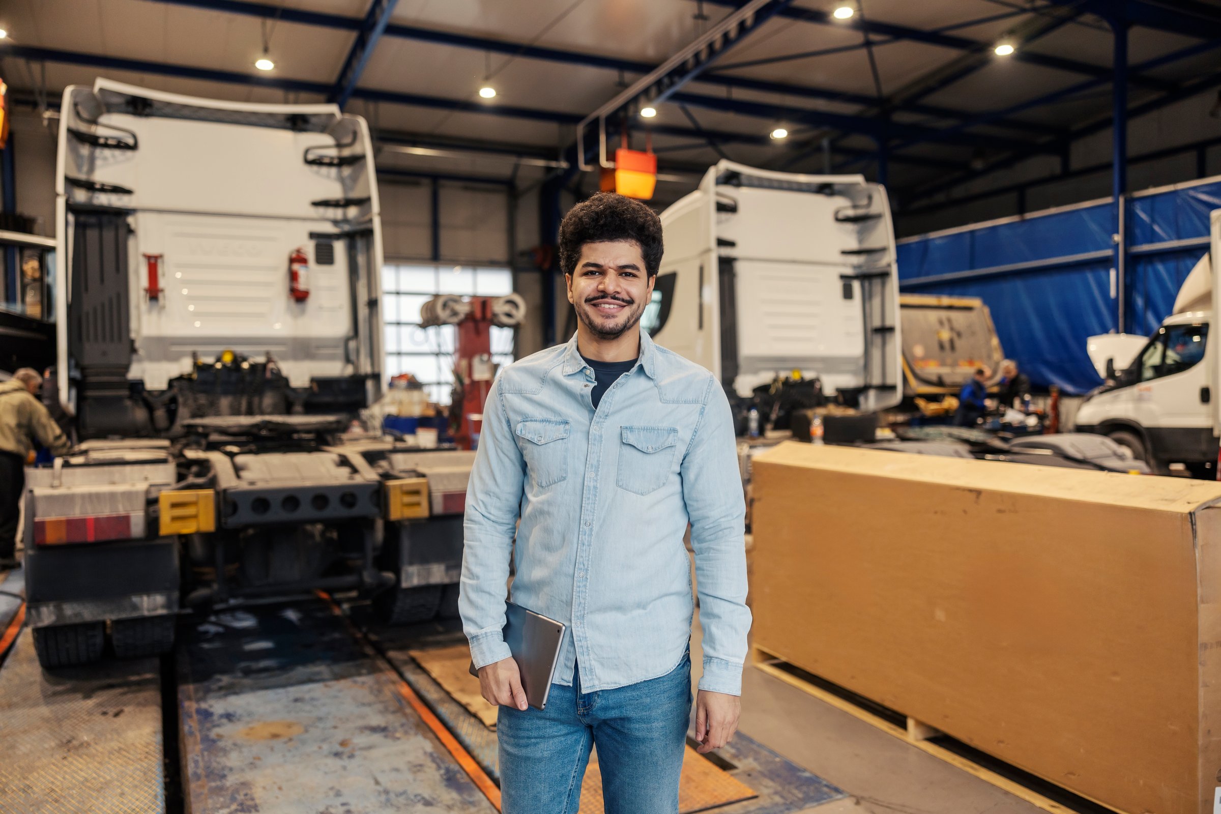 Portrait of diverse logistics company manager with tablet smiling at camera while standing in facility with trucks in blurry background.