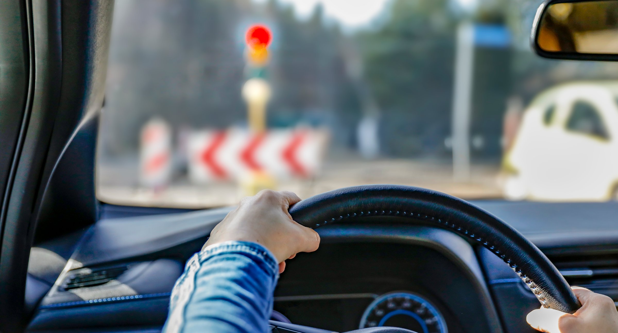Hands holding a car's black steering wheel. The person is wearing a blue sleeve. The vehicle is stopped at a traffic light, visible through the windshield with a red signal illuminated.