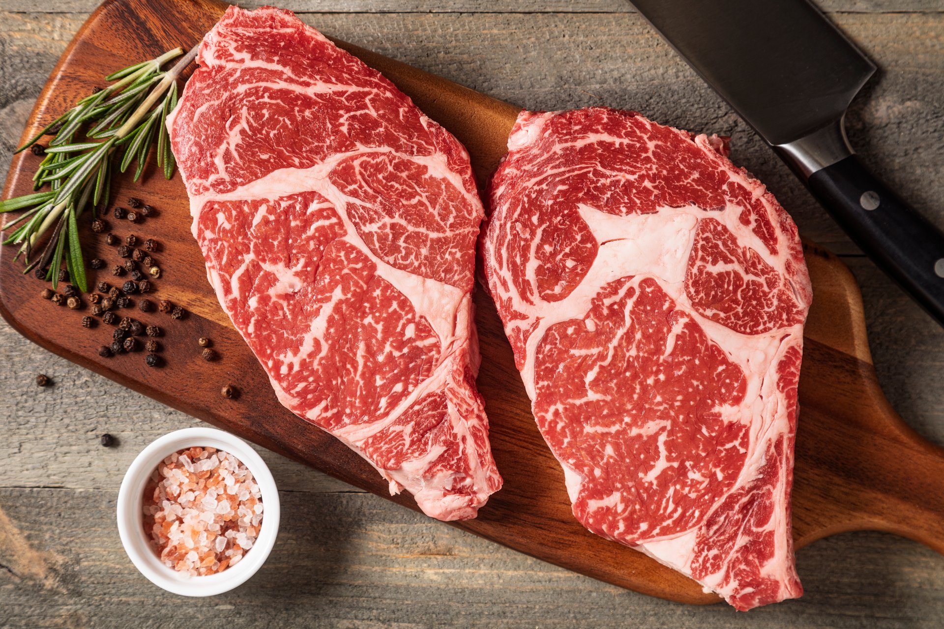 two raw wagyu steaks on a cutting board ready for preparation with knife, rosemary, whole black peppercorns, himalayan pink salt
