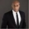 Man in a black suit and tie against a dark background, facing the camera with a serious expression.