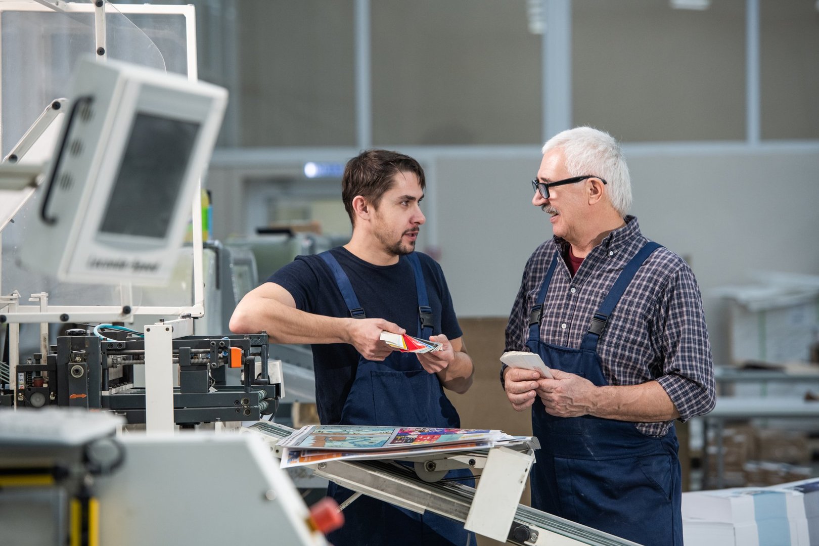 Senior and young workers standing at industrial printer and discussing printing inks while watching color palette