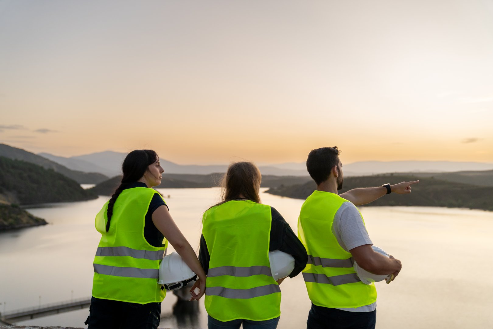Three engineers wearing safety vests are looking at a reservoir at sunset, with one of them pointing towards the water