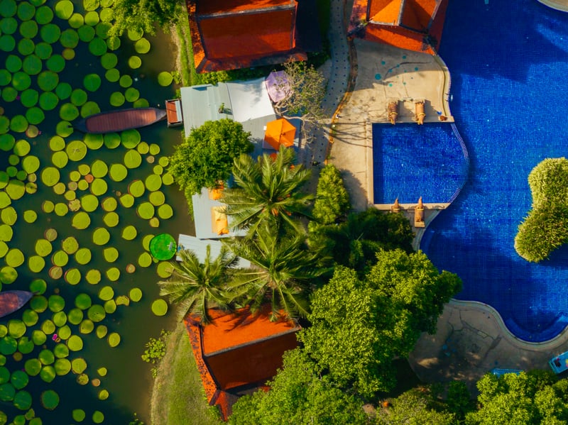 Aerial view of a deep blue swimming pool surrounded by greenery and Thai style buildings, with a lotus pond and wooden boat adding a serene touch.