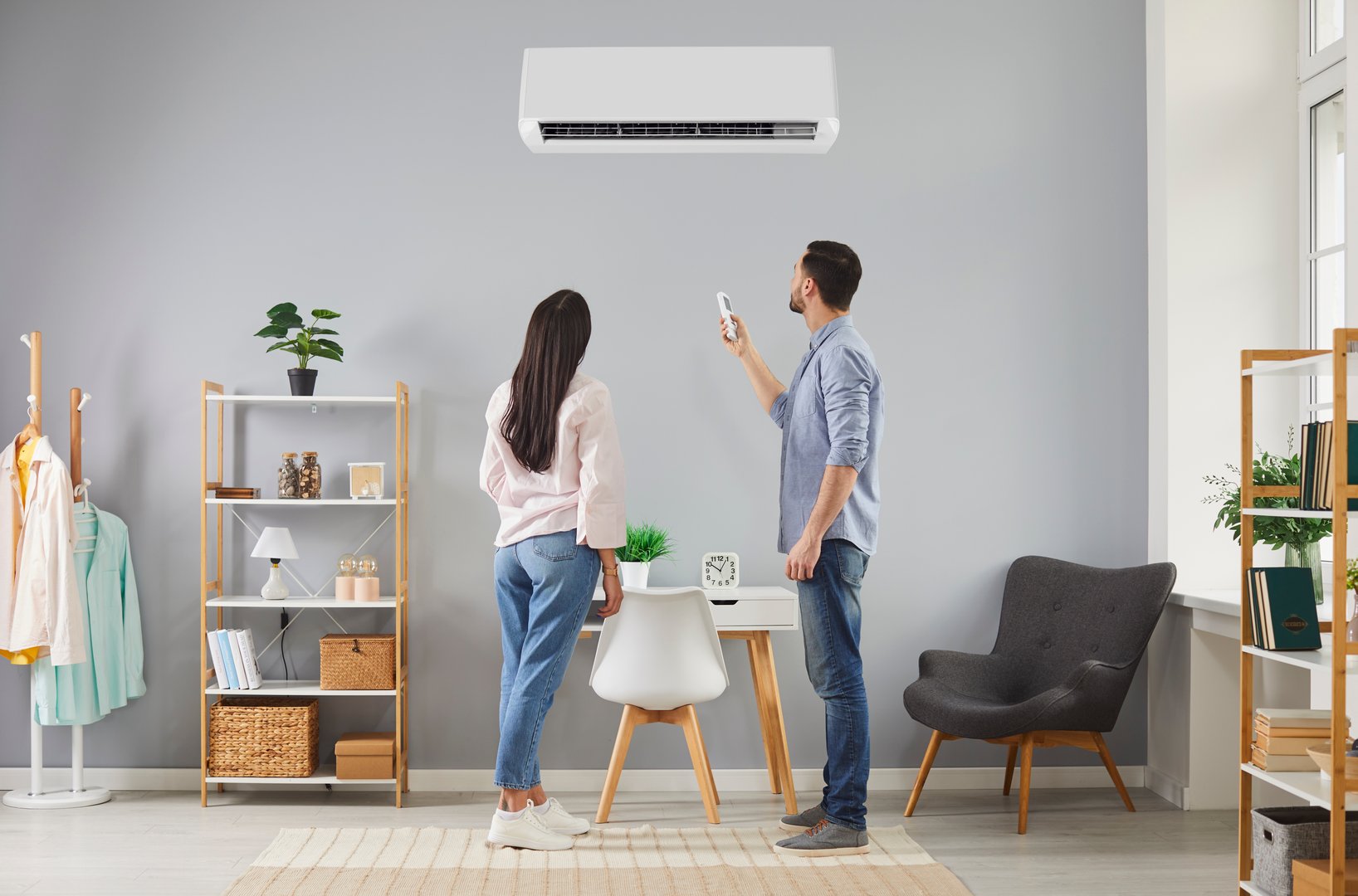 Family couple turn on air conditioning system at home. Young husband and wife standing in living room, looking at white AC unit on grey wall, using remote control and changing temperature in house
