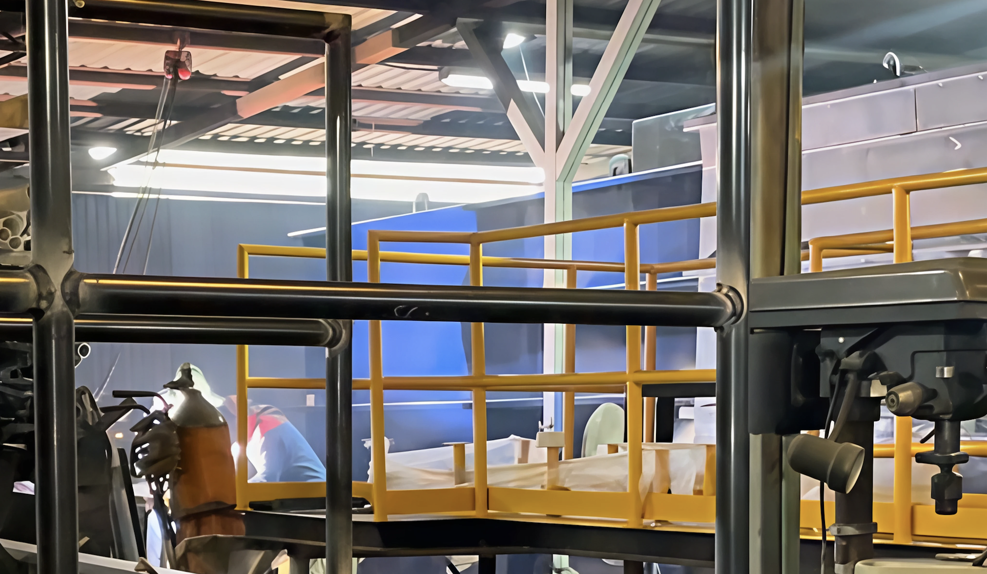 Industrial setting with yellow railings, machinery, and a person working. Metal structures and equipment are visible.