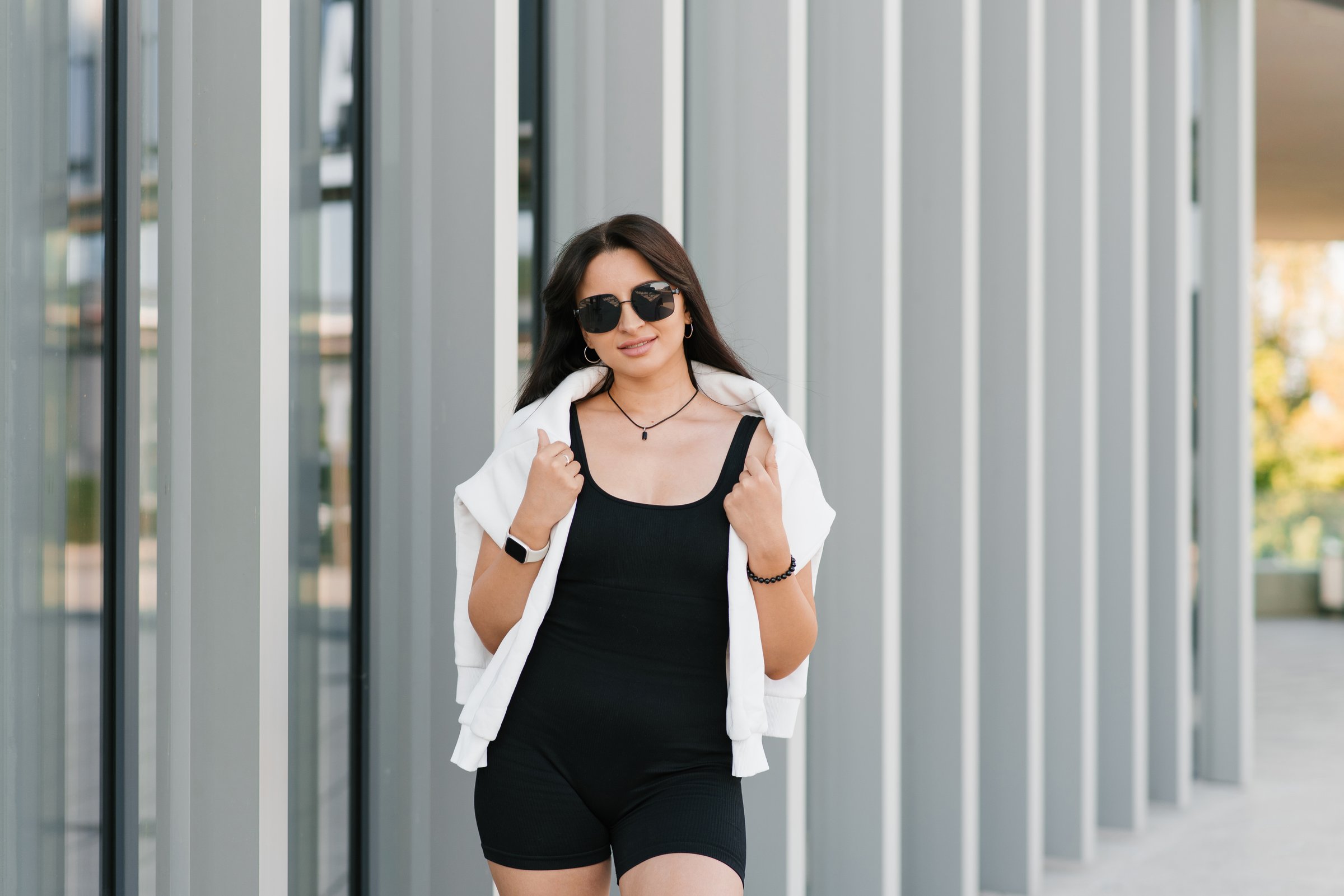 A woman in sportswear walks along a building with large windows. She is wearing sunglasses and a white sweater on her shoulders.
