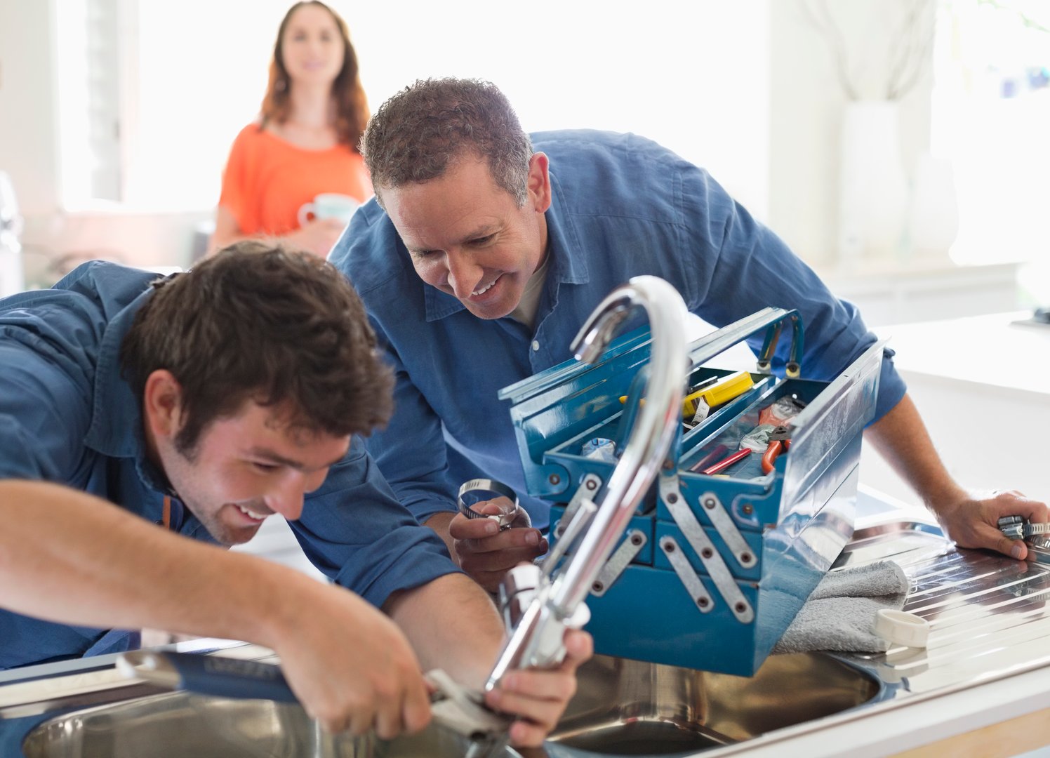 Two men repairing a kitchen faucet with tools; a woman stands in the background, smiling.