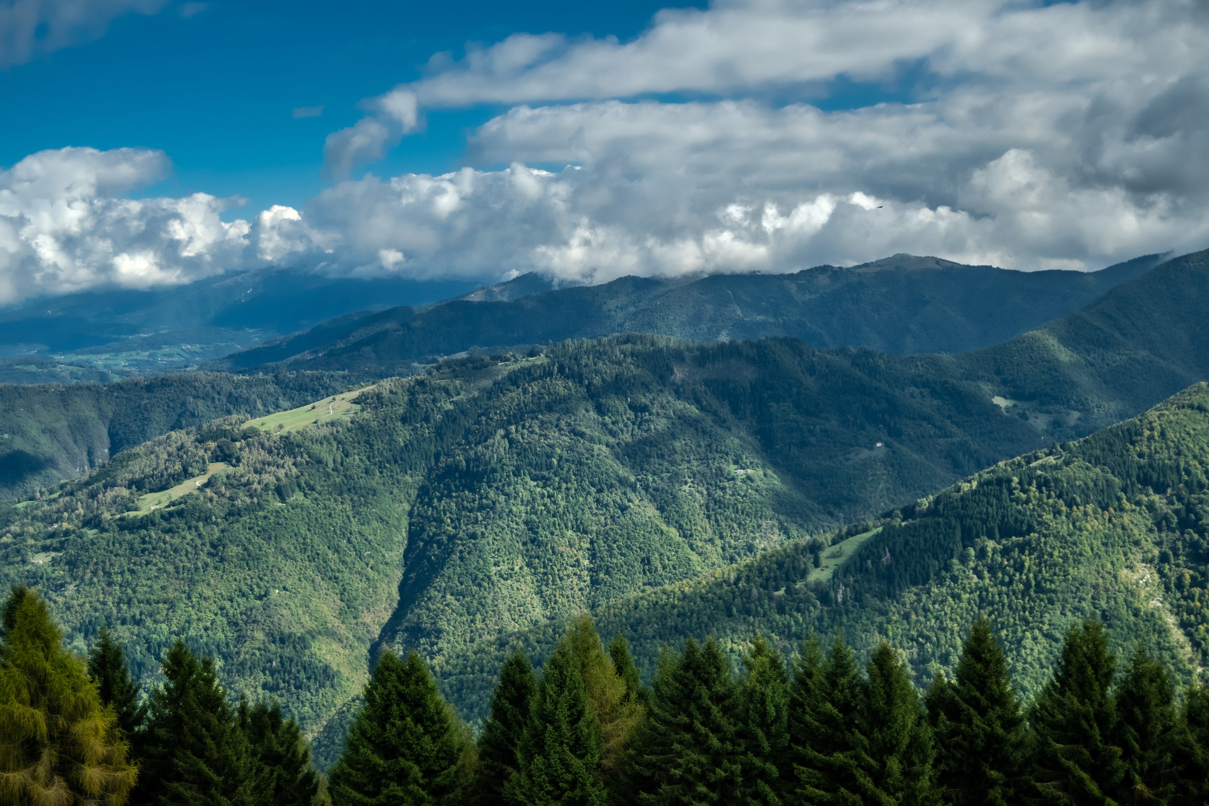 The photo showcases a stunning green mountain range, with lush vegetation cascading down the slopes. Above, a vibrant blue sky stretches endlessly, punctuated by fluffy white clouds that drift lazily, adding depth and contrast to the scene. The interplay of greenery and sky creates a serene and picturesque landscape, inviting viewers to immerse themselves in nature's beauty.