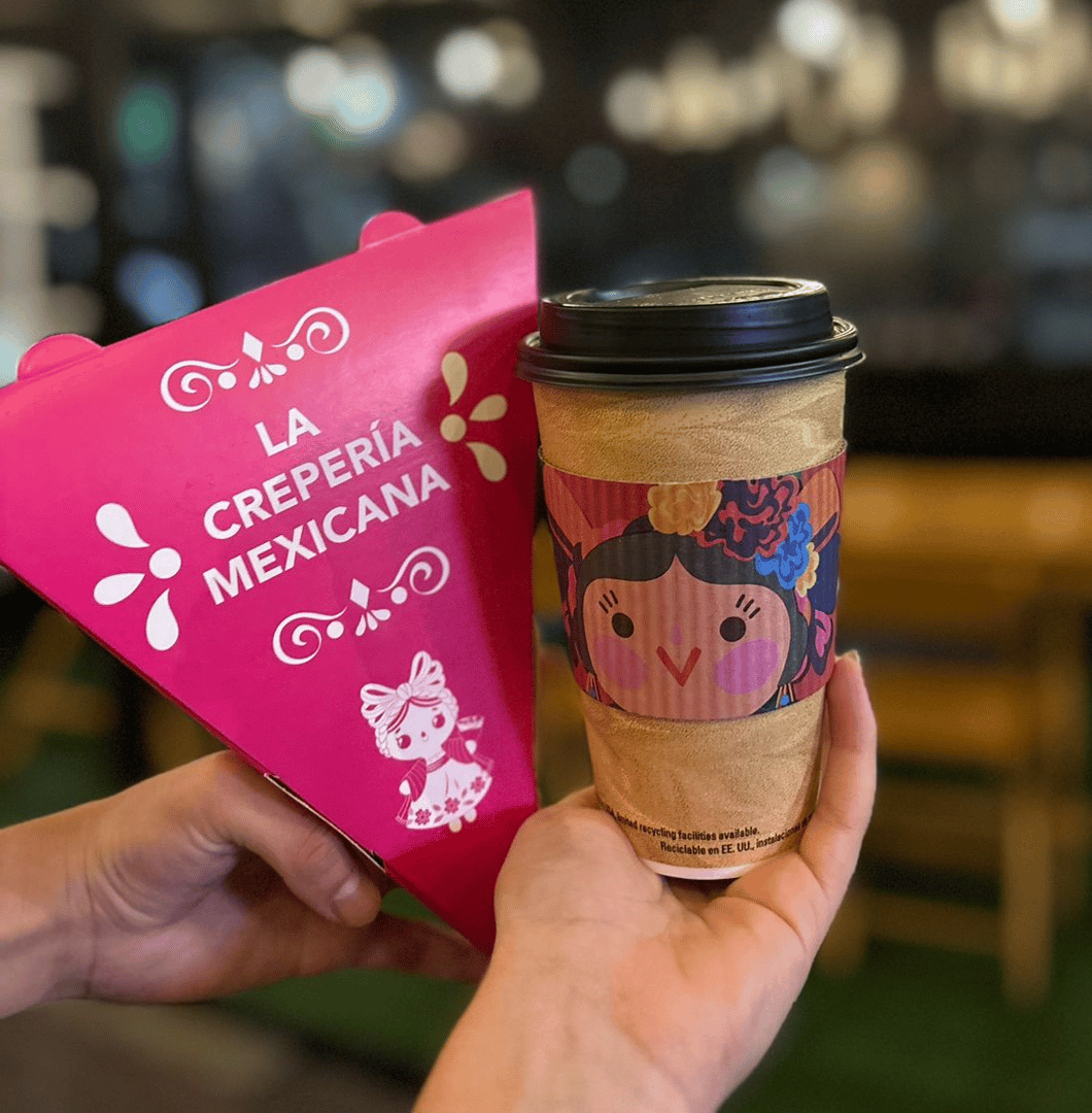 Person holding a coffee cup with a colorful sleeve and a pink box from La Crepería Mexicana, featuring folk art designs.