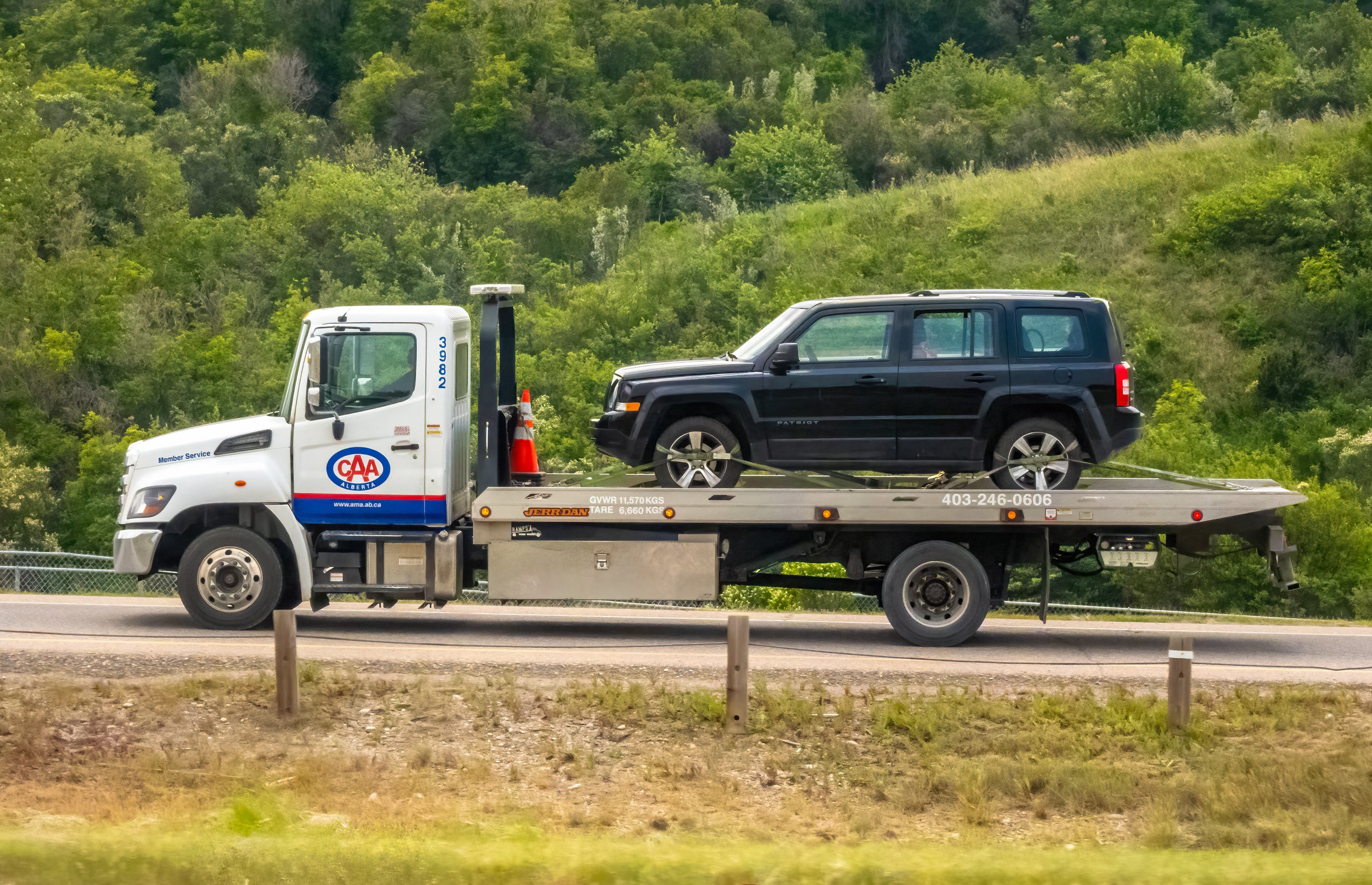 Calgary, Alberta, Canada. Jun 8, 2025. A CAA flatbed tow truck carries a black SUV on a road, framed by lush green hills, symbolizing reliable roadside assistance.